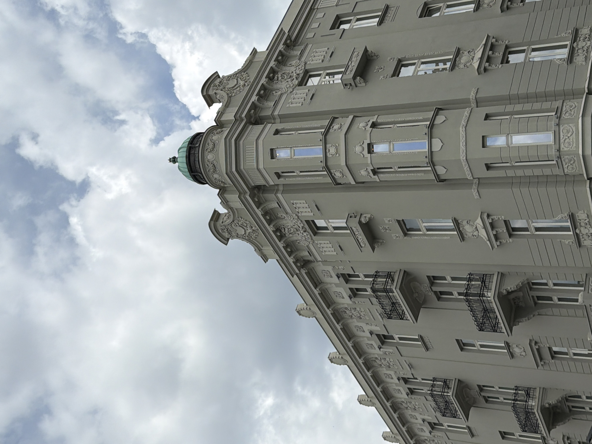 The image depicts a low-angle view of an ornate, multi-story building against a cloudy sky. The building features detailed architectural embellishments, including intricate moldings, cornices, and decorative elements around the windows. Some windows have small balconies with iron railings. The building is painted in a light grey tone. A small green dome sits atop a corner turret. The setting appears to be urban, likely Belgrade, Serbia, based on architectural style, but further evidence would be needed to confirm the precise location. There are no people visible in the shot. The focus is on the architectural features and the contrast between the building and the sky.