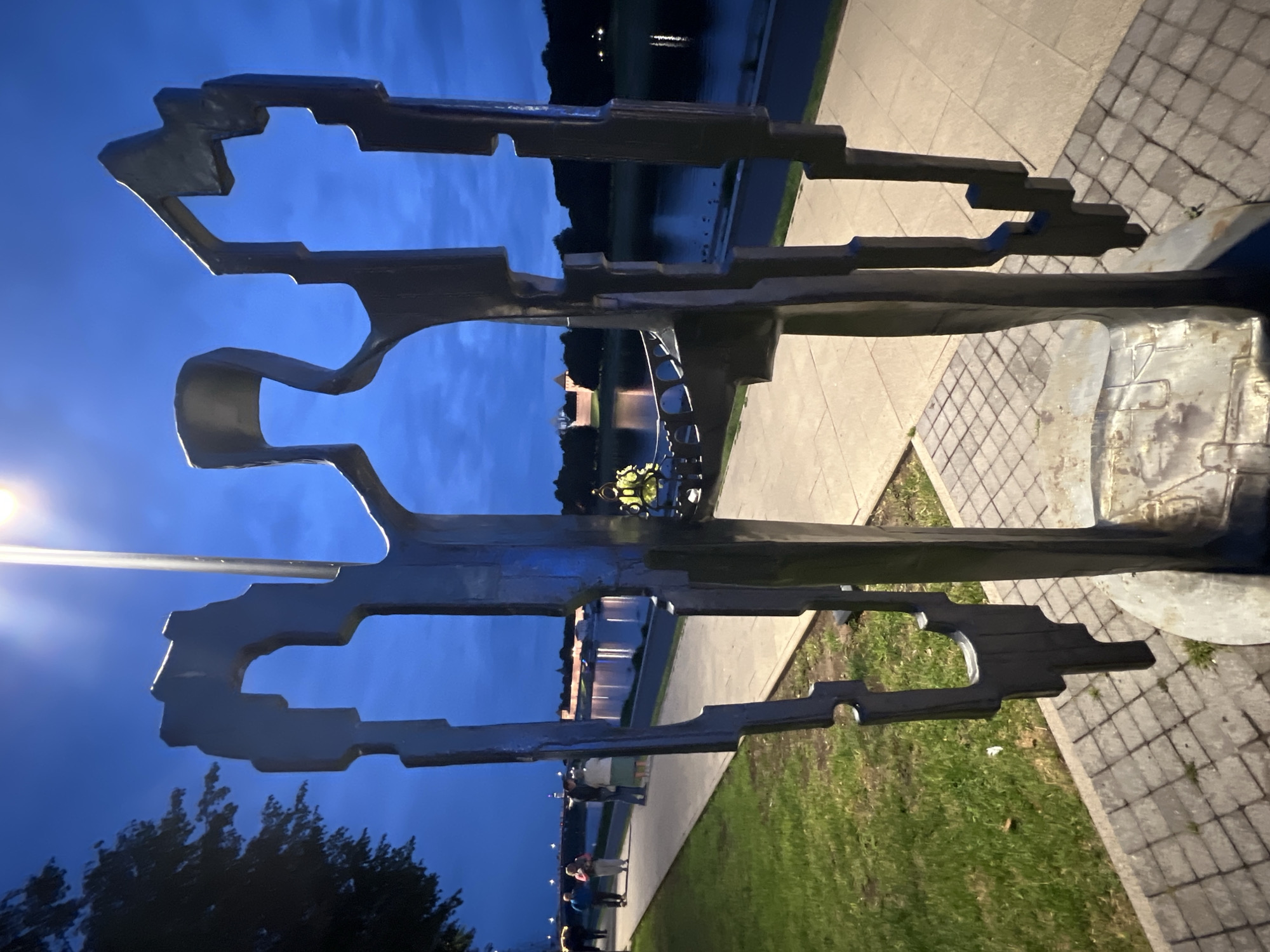 In the city of Veliky Novgorod, Russia, at dusk, a metallic abstract sculpture resembling human figures stands in the foreground. The sculpture is dark and appears to be made of metal, with angular shapes and cutouts. Behind the sculpture, a paved walkway runs alongside a body of water, likely a river, reflecting lights from the opposite bank.
On the walkway, several individuals are visible. To the left, a group of people are walking away from the camera. Further down the walkway, a couple stands talking, and a person in a bright yellow reflective jacket is visible near the water's edge. The background features trees, a distant cityscape with buildings, and a bridge. The sky is a deep blue, characteristic of twilight, with a street lamp emitting a bright light overhead. The ground in the immediate foreground is paved with cobblestones.