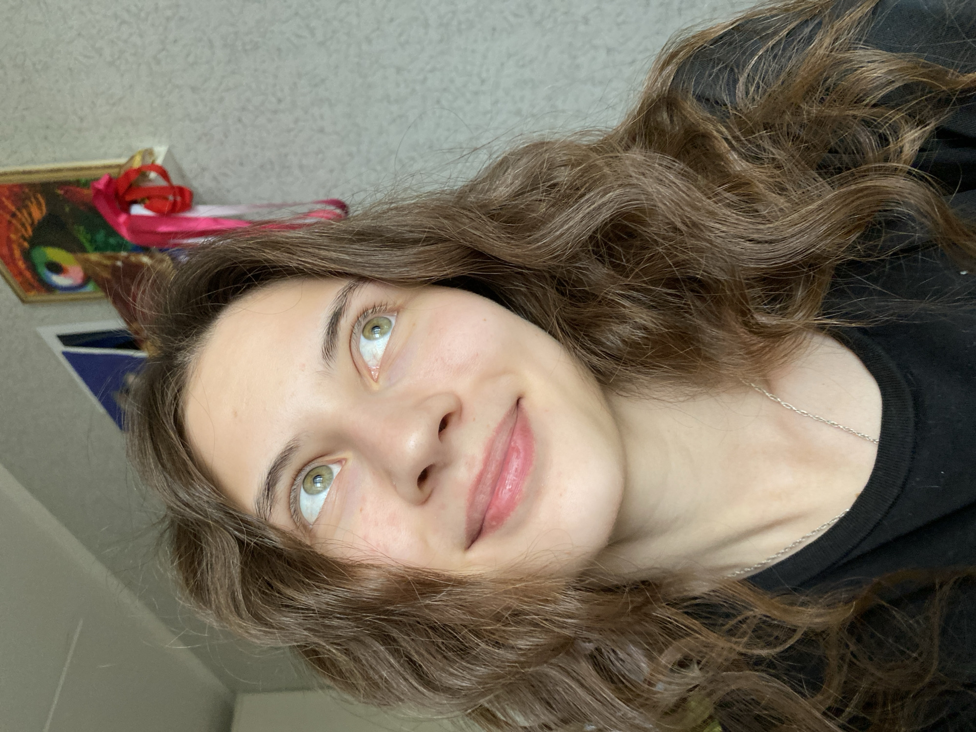 A young woman with wavy brown hair and green eyes is facing the camera. She is wearing a black shirt and a silver necklace. She is smiling slightly and looking up and to the left. The background is blurry and indistinct, showing a wall and a framed painting with red ribbons. There is not enough information to determine if the image was taken in Votkinsk, Russia.