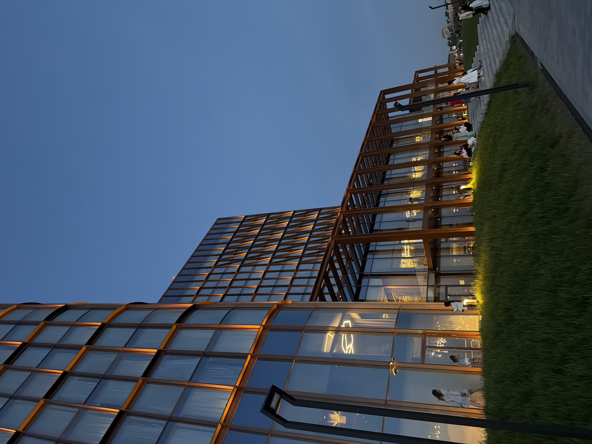 The image presents a low-angle view of modern architectural structures under a twilight sky. The buildings are characterized by their glass facades with grid-like framework. The leftmost building features a curved design, while a taller building sits adjacent to it. An additional structure, with an open framework, is also visible.
Alongside one of the buildings, there is a strip of grass and a walkway or patio where people are situated. The overall atmosphere is one of contemporary urban design.
Location is unknown.