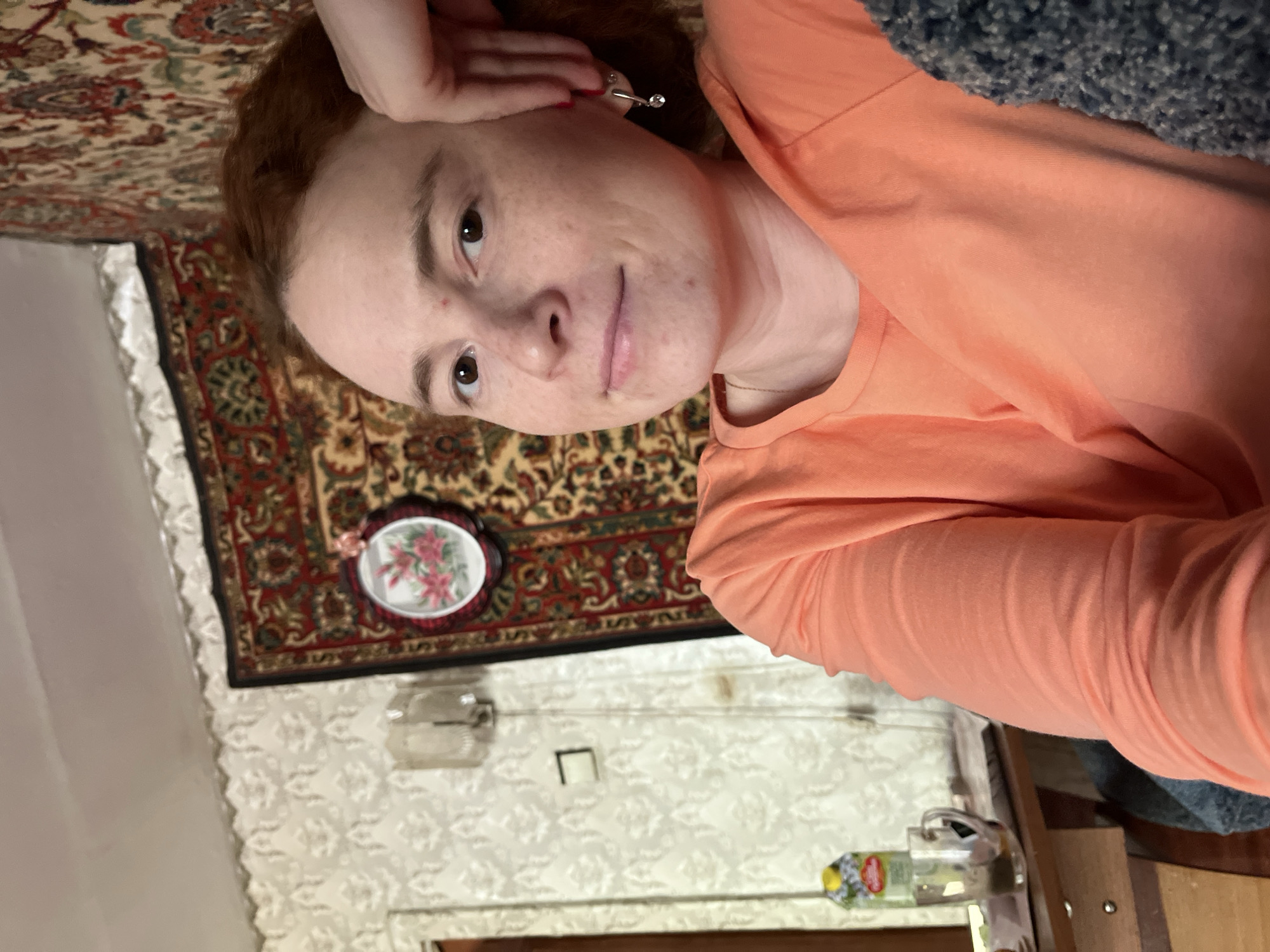 A selfie depicts a woman with light skin and auburn hair, wearing a light orange long-sleeved shirt. She is lying down, with her head resting on her hand and an earring visible in her left ear. Her expression is a slight, closed-lip smile.
Behind her, the environment suggests an interior setting. There is a patterned carpet on the wall. Below the carpet, the wall is decorated with patterned wallpaper and a small, framed floral picture. A table with miscellaneous items, including a bottle, is visible in the background.
The location is Chaikovsky, Russia.