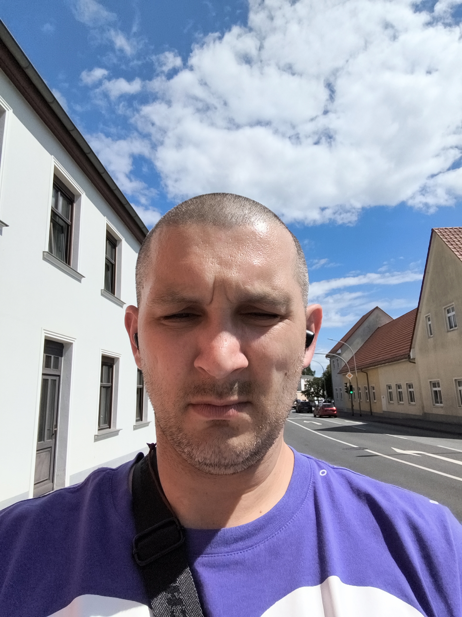 A man takes a selfie while standing on a street in Wolgast, Germany. He is wearing a purple t-shirt and has a black strap across his chest, likely from a bag. He has a buzz cut and is wearing an earbud in his right ear. The background shows a white building on the left and houses with red roofs on the right. A street with cars and a traffic light is visible in the distance. The sky is blue with white clouds.