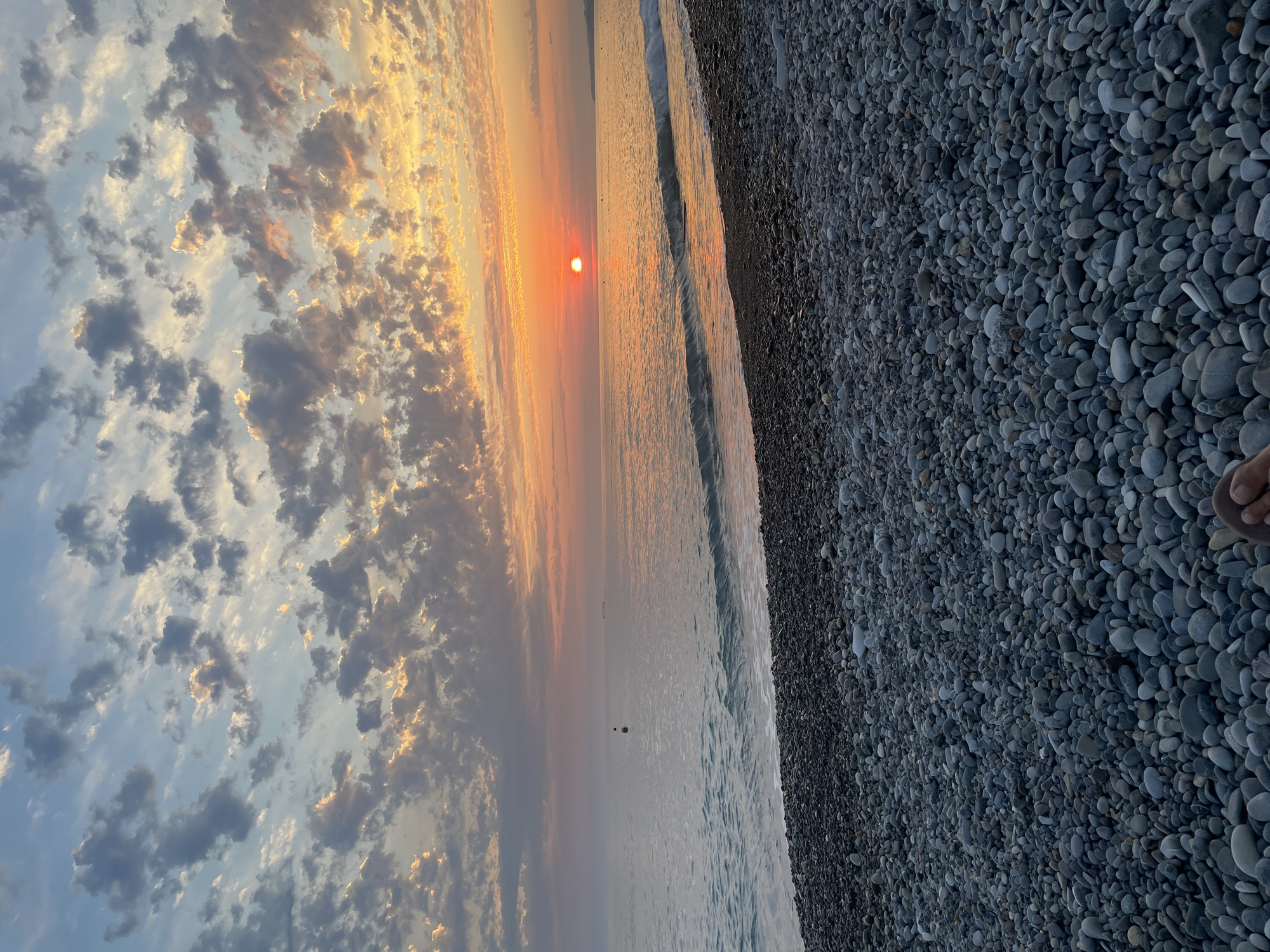 This is a full shot photograph of a pebble beach with the ocean and a sunset in the background. The foreground is dominated by a dense field of smooth, gray pebbles of various sizes. The pebbles are wet and appear to be recently washed by waves. In the middle ground, the gentle waves of the ocean lap onto the shore. A small, dark buoy is visible in the water to the left of the frame, and a distant ship is on the horizon to the right. The background features a vibrant sunset with the sun, a bright orange orb, low in the sky, casting a golden reflection on the water. The sky is filled with scattered clouds, some tinged with orange and yellow from the sunset. The bottom right corner of the image shows the bare feet of a person partially visible, resting on the pebbles, suggesting a relaxed and observational pose. The overall scene evokes a sense of tranquility and natural beauty.
