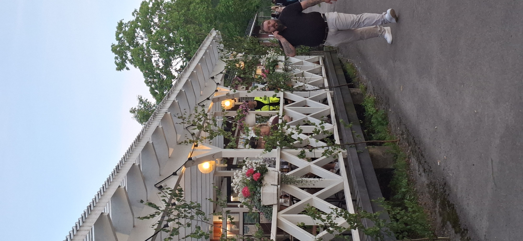 The image depicts an outdoor seating area of a restaurant or cafe, likely in the city of Salpakangas, Finland. The establishment features a white wooden gazebo with decorative latticework and hanging plants, illuminated by string lights. Several people are seated at tables under the gazebo, appearing to be patrons. A man in a black polo shirt and light-colored pants is walking on a paved path in the foreground, talking on his mobile phone. He has tattoos visible on his arm. Behind the gazebo, a red building and lush green trees are visible, suggesting a natural or park-like setting. The sky is clear and blue. The overall atmosphere appears casual and relaxed.