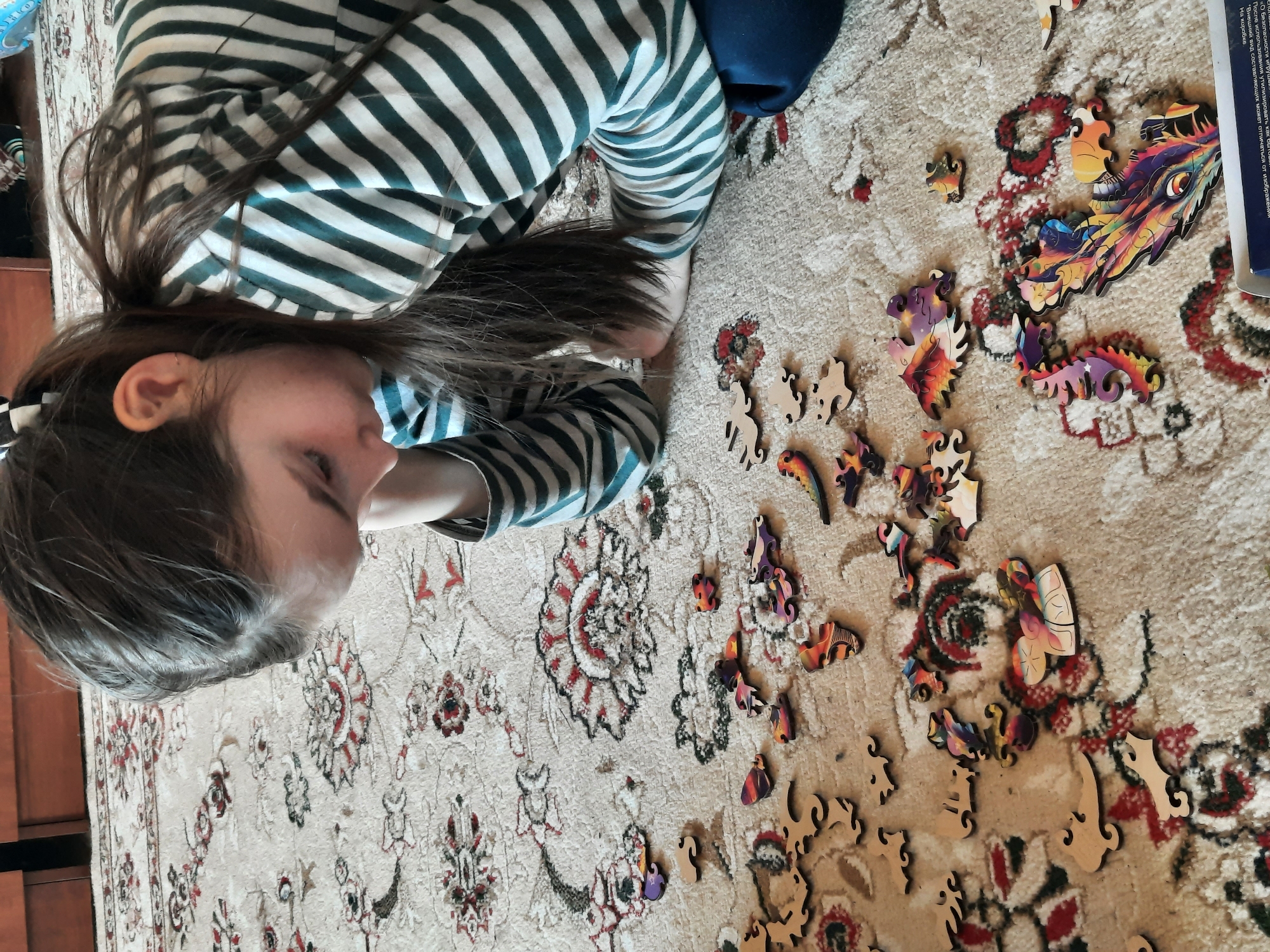 A young person with long brown hair and wearing a striped shirt is lying on a patterned rug, concentrating on assembling a wooden puzzle with colorful pieces scattered around them. The rug features a floral design with red and beige tones. A puzzle box is partially visible in the background, along with text in Russian indicating it is about the safety of toys. The photo was taken in Russia.