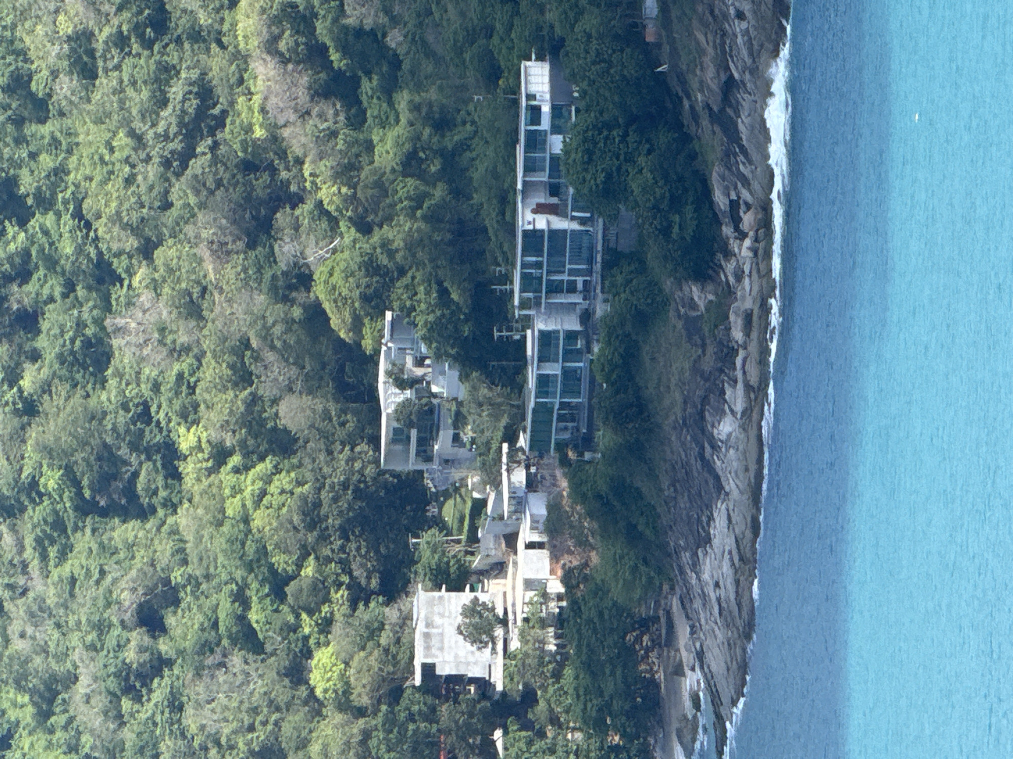 The image shows a cluster of modern villas nestled into a lush, green hillside overlooking a rocky coastline and bright turquoise water. The villas are predominantly white, with large glass windows and clean, geometric lines. Some appear to be under construction, with exposed concrete and scaffolding. The natural environment is characterized by dense, tropical foliage covering the steep slopes. The sea is calm with gentle waves lapping against the shore. This scene is located in Rawai, Thailand. There are no people visible in the image.