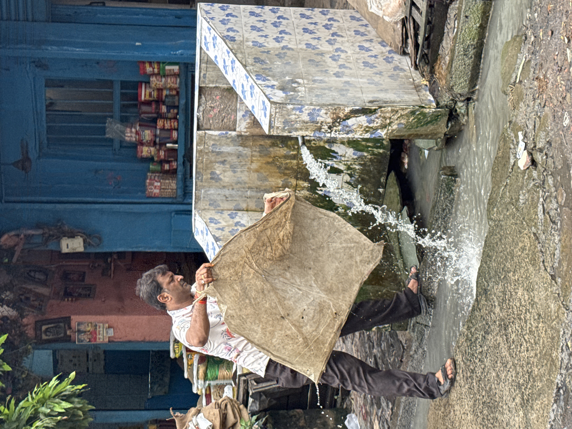 In an outdoor scene, possibly in a marketplace or residential area of Kolkata, India, a man is washing a large, tan-colored piece of fabric near a tiled structure with a water outlet. The fabric appears to be old and somewhat dirty, with visible holes and stains. The man, wearing a short-sleeved patterned shirt and dark pants, is holding the fabric under a stream of water gushing from a pipe that emerges from the tiled structure. The tiled structure is decorated with blue floral patterns and shows signs of age and wear, with some discoloration and moss growth. A small channel or gutter runs in front of the structure, carrying away the water. In the background, there are buildings with blue and pink facades, and shelves stocked with various goods, suggesting a commercial setting. The ground is wet and uneven, with some stones and debris visible. The overall impression is of a utilitarian task being performed in a busy, possibly developing, urban environment.