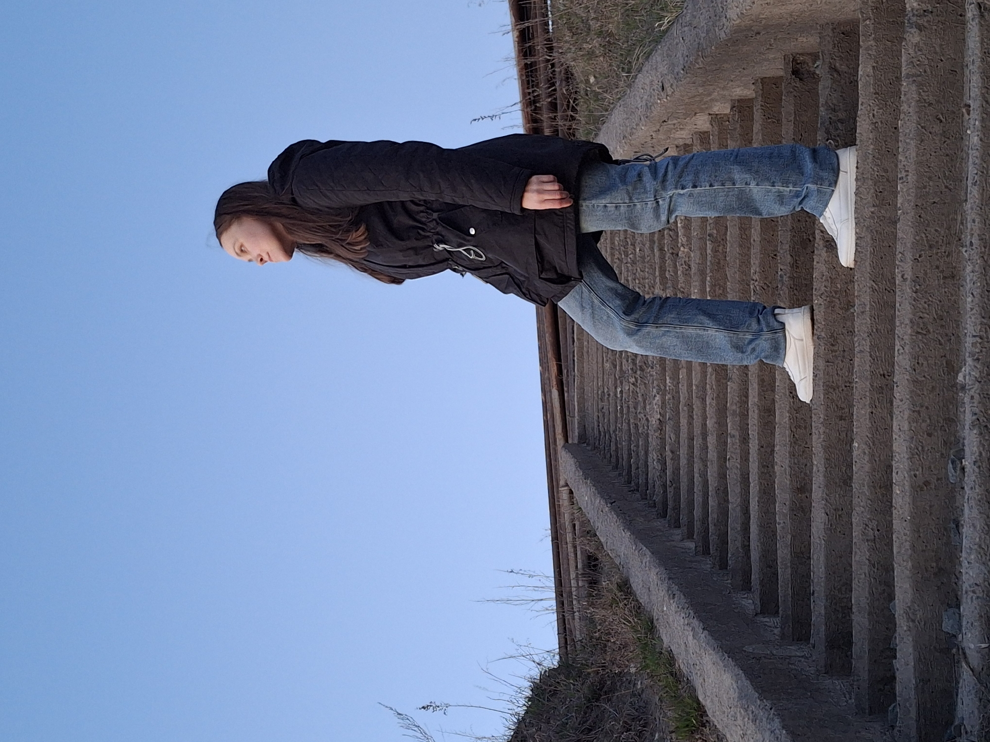 A young woman with long brown hair, wearing a black puffer jacket, blue jeans, and white sneakers, is lying on a flight of concrete stairs. The stairs are weathered and appear to be part of a structure in an outdoor environment. The background includes a clear blue sky. The location is likely in Chaikovsky, Russia.