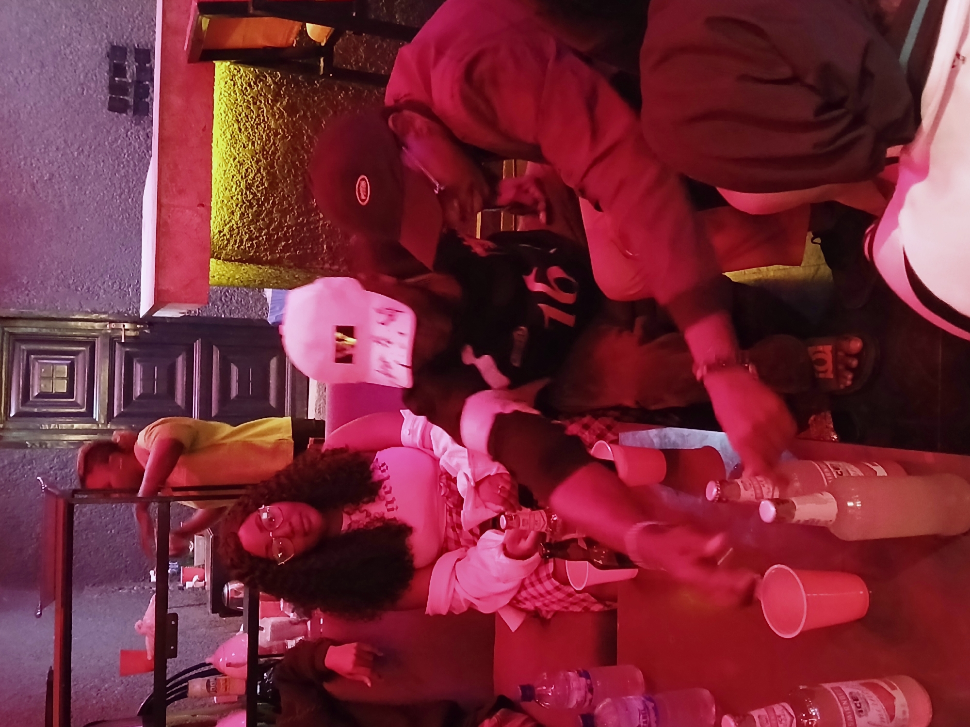 The image depicts a gathering in what appears to be a bar or lounge setting on Rukuba Road, Nigeria. Several individuals are present, with one woman lying down on a surface covered with a checkered cloth, bottles, and plastic cups. A man wearing a baseball cap leans over her, possibly pouring a drink. Another person lies down on a nearby surface. The environment is illuminated with a red light, which gives the scene an overall reddish tone. In the background, a door with square patterns can be seen. Additional people are standing around the table, visible from their torsos and arms.