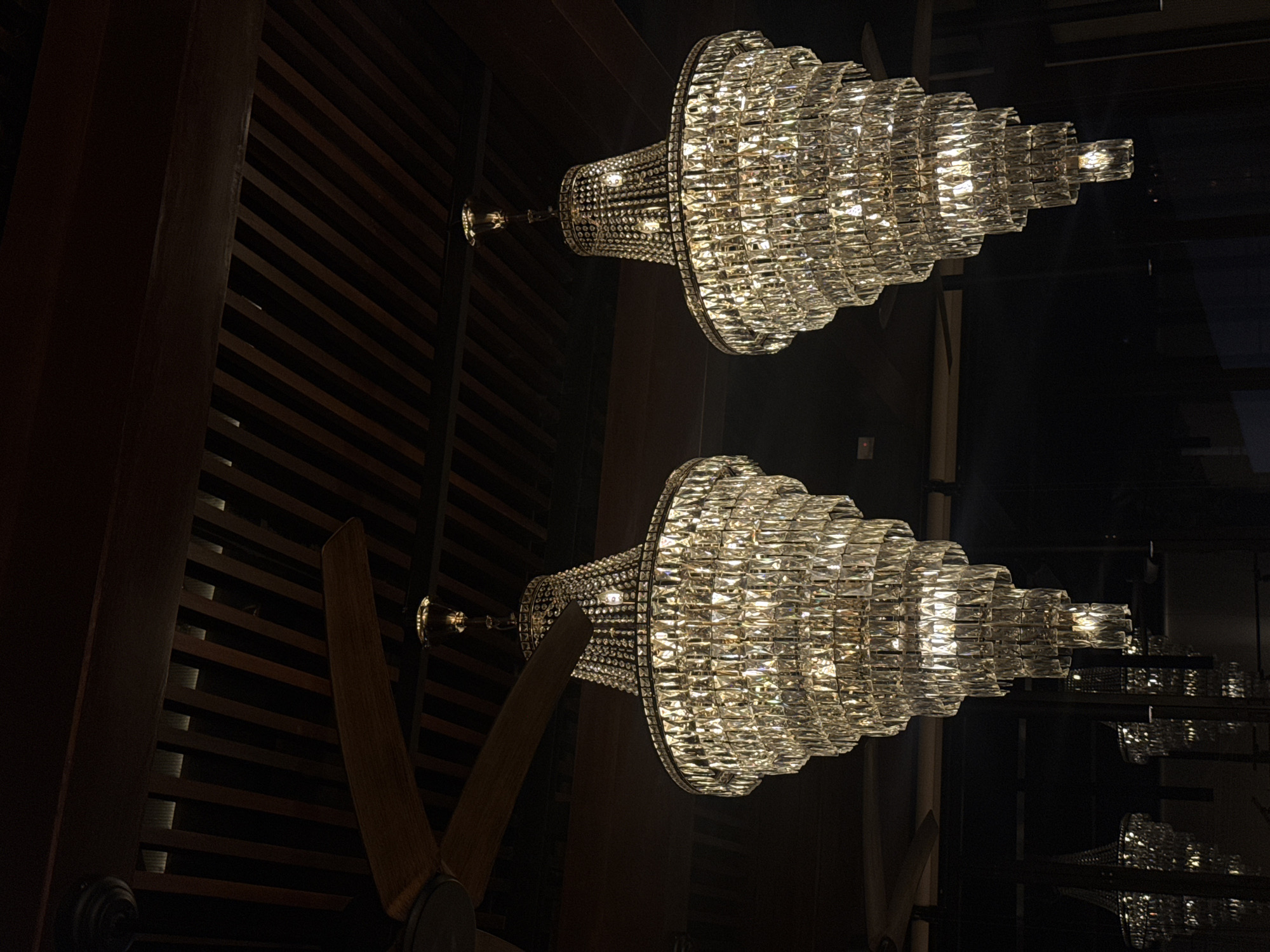 The image displays two large, ornate crystal chandeliers illuminated against a dark background, oriented diagonally across the frame as if hanging horizontally. Each chandelier is tiered, formed from numerous rectangular crystal elements that sparkle brightly, emitting a warm, golden light.
The background to the left features dark wooden paneling with horizontal slats, suggesting a textured wall or ceiling. A dark, broad wooden beam or column is partially visible along the far left edge of the image.
In the lower left portion of the image, parts of a ceiling fan, including its wooden blades and base, are visible beneath and beside the lower chandelier.
The overall scene is dimly lit except for the radiant chandeliers, indicating an indoor setting, likely a grand interior such as a hotel lobby, restaurant, or event hall, perhaps during the evening or night. The perspective is from below, looking upwards at the light fixtures. No people or specific activities are depicted, and no text is visible.