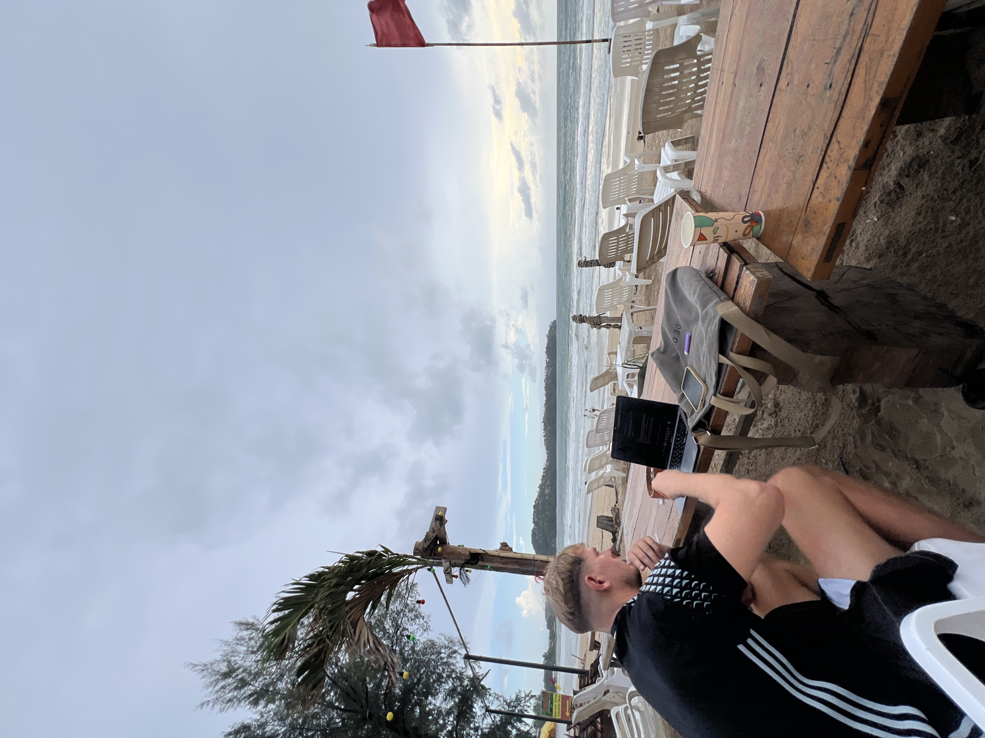 The photograph shows a man sitting at a wooden table on a beach in Unknown, Unknown, working on a laptop. The beach is lined with white plastic chairs. A red flag is visible in the background near the ocean. A palm tree and other foliage are in the foreground. The sky is overcast.