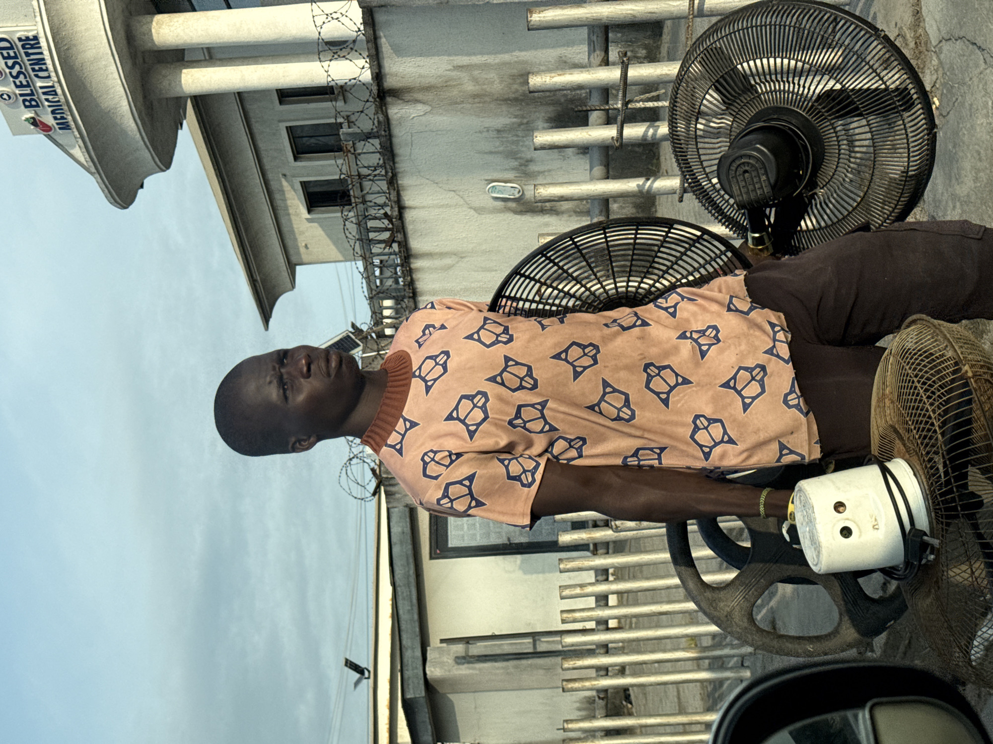 A person stands in front of the Blessed Medical Centre in Igboefon, Nigeria. The person is wearing a light brown shirt with a repeating pattern of blue geometric shapes and dark brown pants. The person is holding a white appliance with two visible circular openings and a black power cord. Behind the person, two black oscillating fans are visible, one to the left and one to the right, positioned on top of each other. The medical center is a white building with large pillars and a curved roof. A gray concrete wall topped with barbed wire separates the street from the medical center. The ground is paved with concrete. The sky is clear with a few wispy clouds.