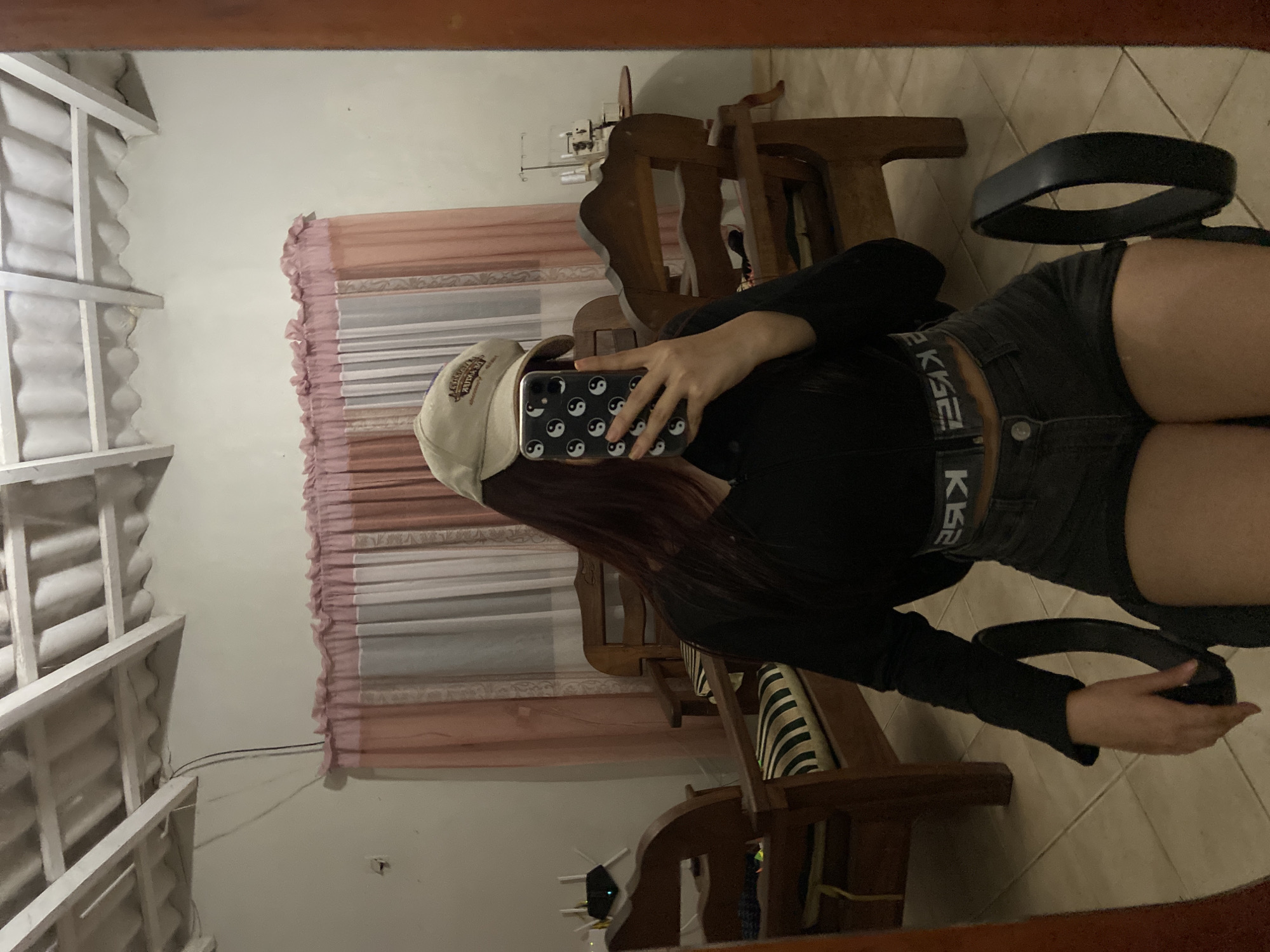 A person wearing a baseball cap, black long-sleeve shirt, and dark shorts is taking a selfie in a room with wooden furniture and a tiled floor. The phone has a black case with yin and yang symbols. A black belt is in the person's hand. The room has white walls, pink curtains, and a visible white slatted ceiling. The image appears to be a reflection in a mirror, and the location details (Juan Griego, Venezuela) are not visually ascertainable from the photo.