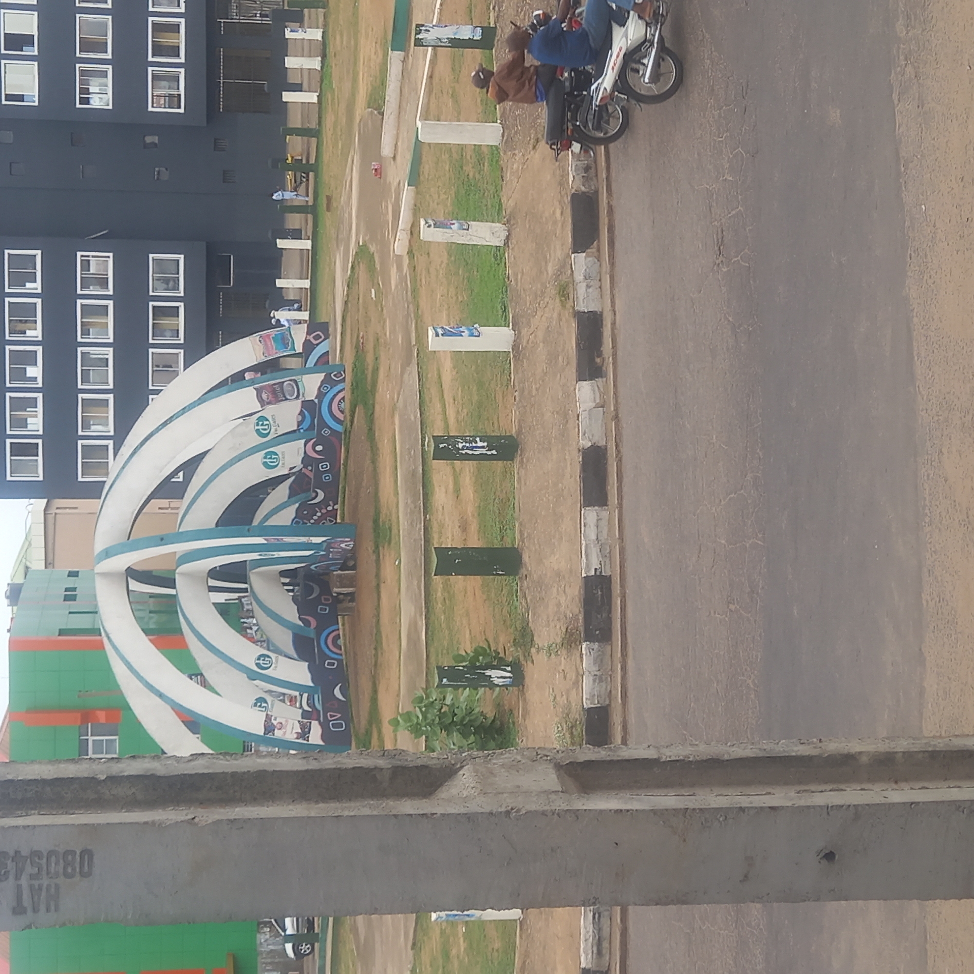 The scene depicts a street-level view in Dadi Riba, Nigeria. The foreground is dominated by a concrete structure, possibly part of a building under construction. The middle ground features a curved, white-and-blue archway structure adorned with various designs. Adjacent to it is a grassy area punctuated by white and green pillars. A motorcycle with two riders is visible on the paved road. In the background, buildings with green and orange, as well as dark blue with white windows are visible.
