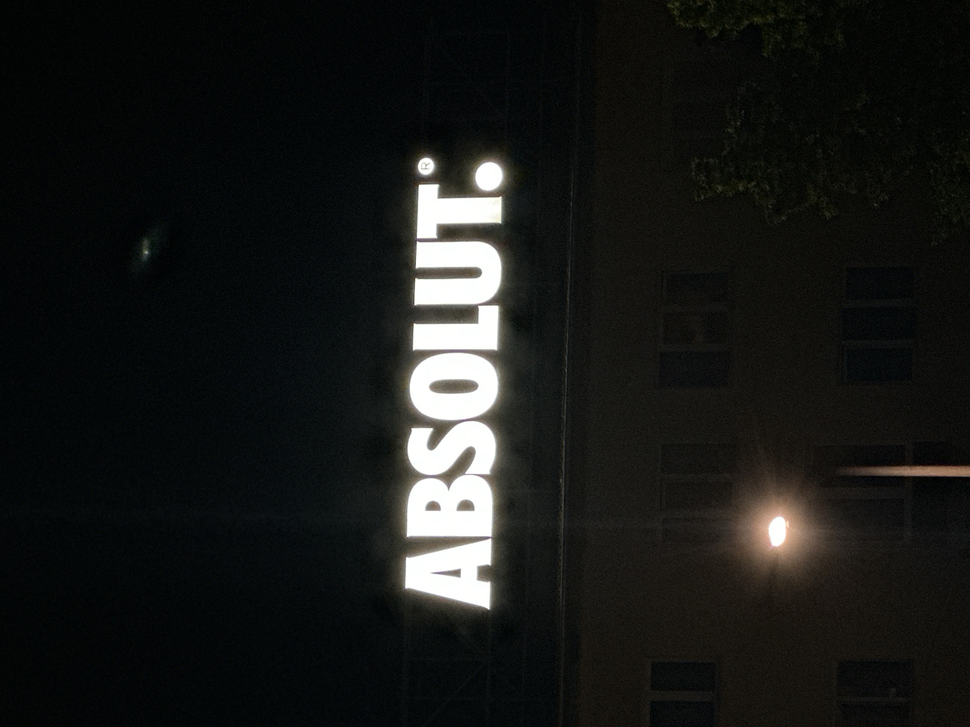 The image is a nighttime shot of a building with a prominent lit sign that reads "ABSOLUT:". The sign is white and illuminated, positioned above the building. Below the sign, the building appears to be a multi-story structure with several windows visible. One street lamp is lit, casting a warm glow. To the right of the building, some trees are visible, their foliage dark against the night sky. The overall scene suggests an urban environment at night.
The sign is located on the building at 1-3, Boulevard Arsenalski, in Sofia, Bulgaria. This location is part of a commercial area in the city.