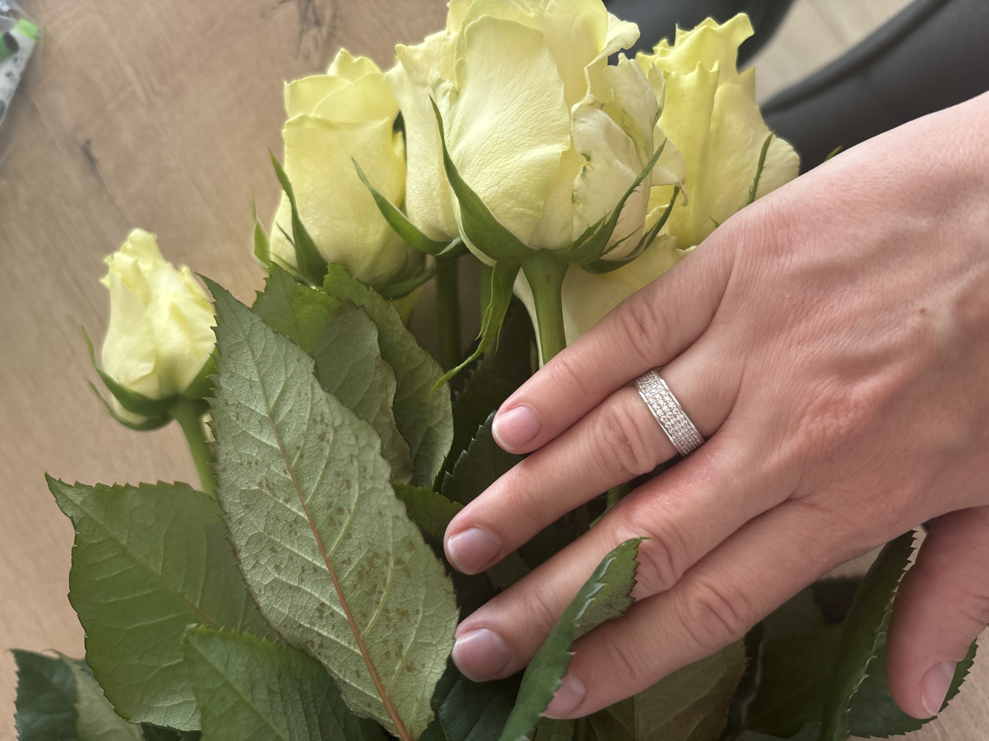A close-up shows a hand with a diamond ring resting amongst a bouquet of pale yellow roses with green foliage. The hand appears to be gently touching the leaves. The roses are in various stages of bloom, from closed buds to partially open flowers. The background is a light brown surface, possibly a wooden table. There is a dark object in the upper right corner, possibly a chair or piece of furniture. There is no evidence to suggest the location is Chaikovsky, Russia.