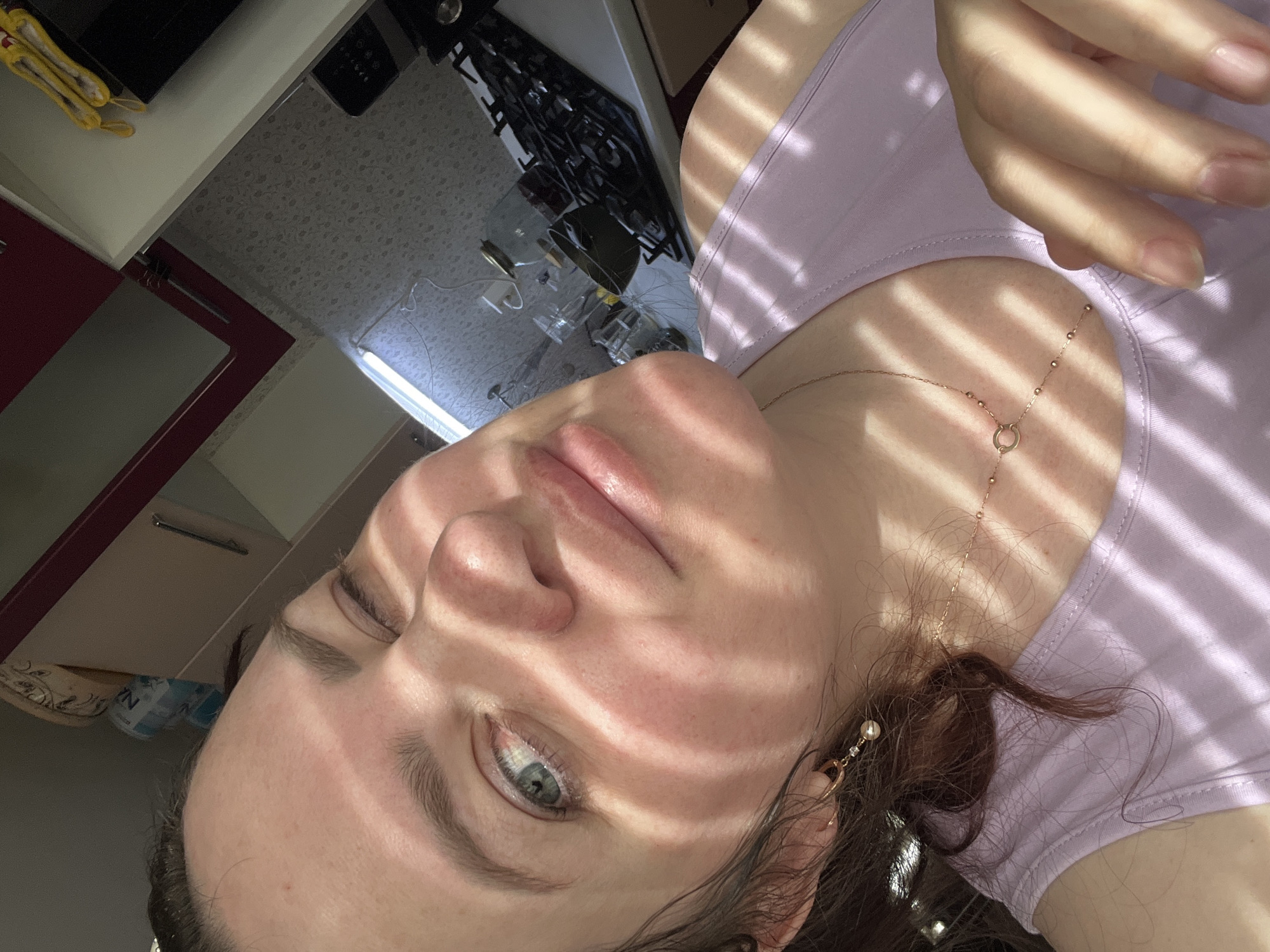 A close-up, angled self-portrait of a woman, possibly in her late teens or early twenties, is presented. The subject is indoors, likely in a kitchen, given the presence of countertops, cabinets, and a stovetop visible in the background. She is wearing a light purple top and a delicate, gold-colored necklace. The scene is illuminated by sunlight filtering through blinds or louvers, casting stripes of light and shadow across her face and clothing. Her facial expression is neutral, with a slightly relaxed gaze. She is wearing an earring on her right ear. The location is not Noviy, Russia; this cannot be determined from the image.