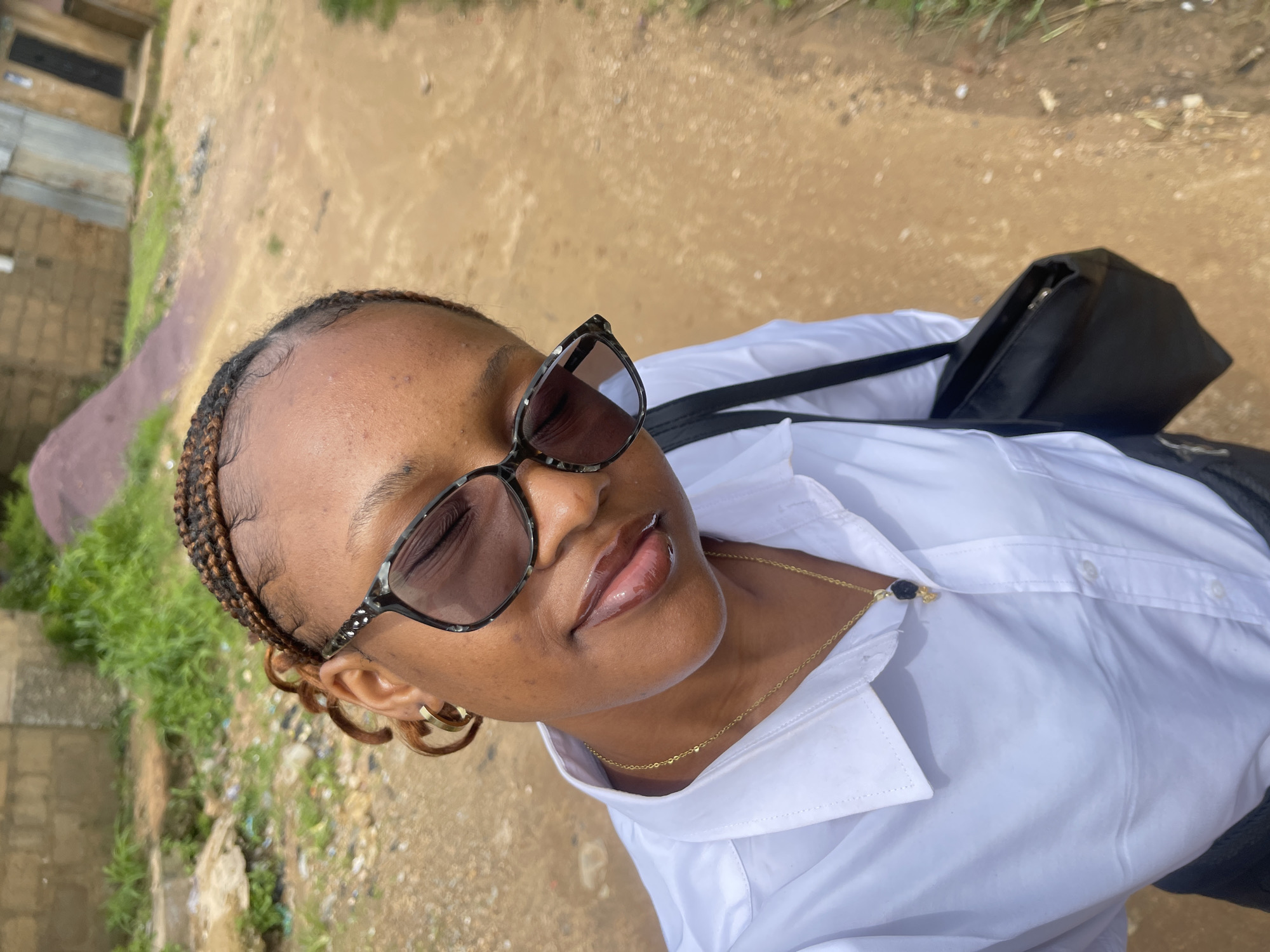 A young Black woman with braided hair and sunglasses takes a selfie outdoors, likely in Rafin Par, Nigeria, given the context. She is wearing a white collared shirt and a gold chain with a dark pendant. A black bag is slung over her shoulder. The background shows a dirt path, overgrown grass, and rudimentary buildings, characteristic of a rural or semi-urban setting in Nigeria. The lighting suggests it is daytime.
