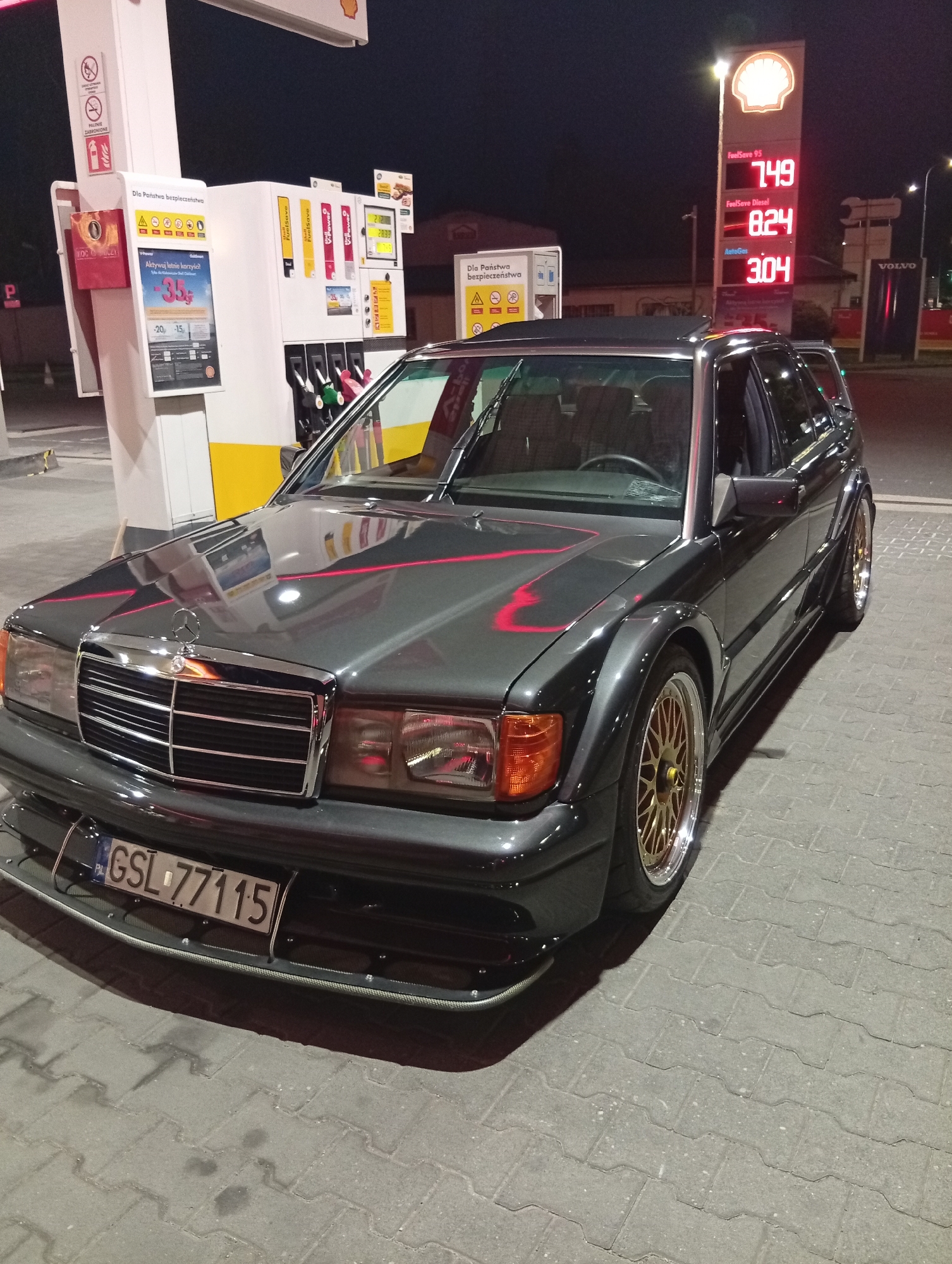 The image captures a custom dark gray Mercedes-Benz 190 E 2.3-16 Cosworth wagon parked at a Shell gas station at night. The car features gold multi-spoke wheels and red racing stripes on the hood and fender. The gas station's illuminated price board is visible, displaying prices for FuelSave 95 at 7.49, FuelSave Diesel at 8.24, and AutoGas at 3.04. A white and yellow gas pump structure is to the left of the car, with safety warnings and promotional materials displayed. In the background, a Volvo dealership sign is visible, along with streetlights and buildings. The ground is paved with interlocking gray bricks. The location is identified as Wrocław, Poland. There are no people visible in the image.