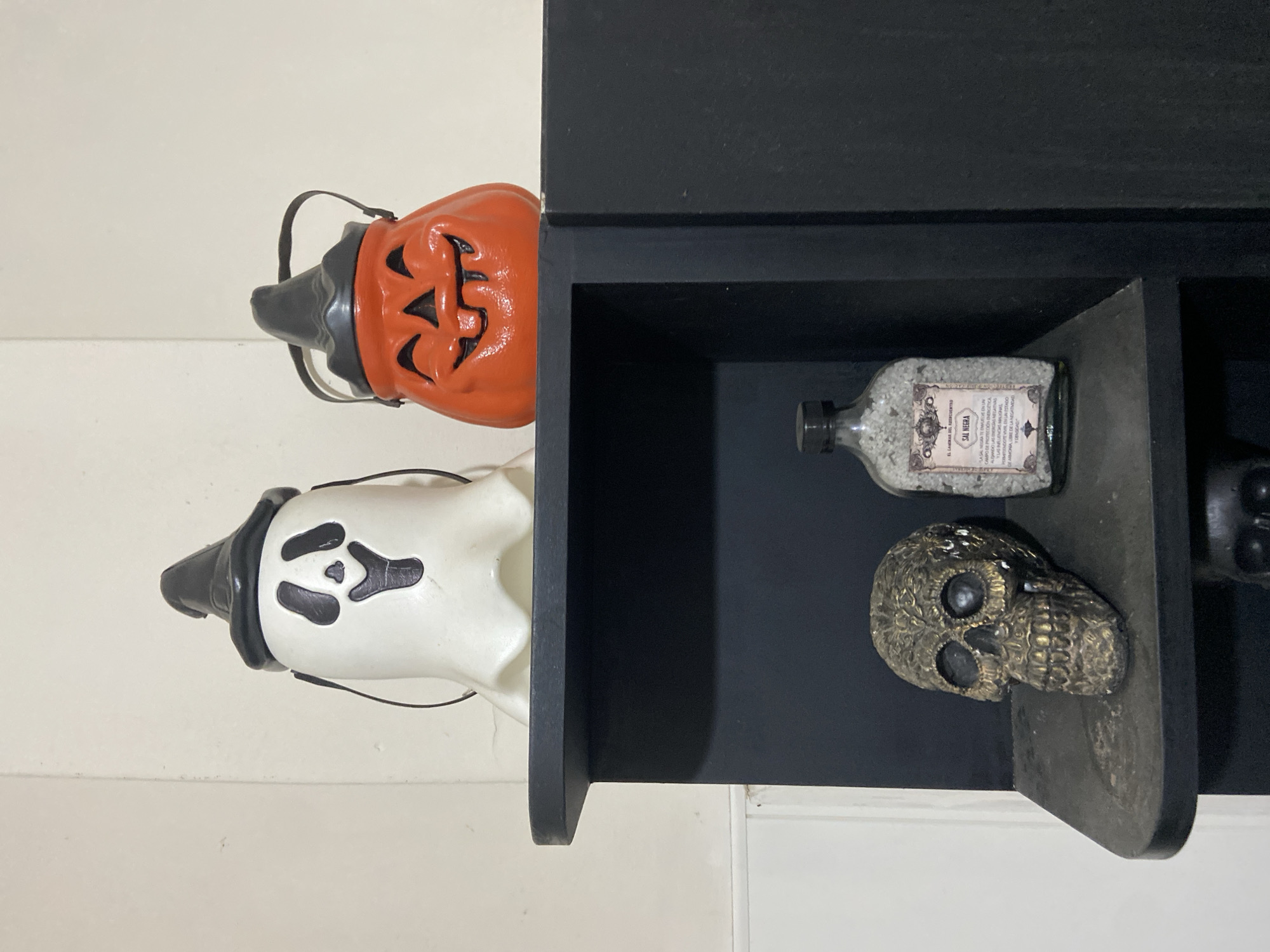 This image captures an indoor scene in Buenos Aires, Argentina, featuring Halloween-themed decorations and esoteric items displayed on a dark wooden shelf.
On the left side, two classic Halloween treat buckets are visible. The top one is an orange jack-o'-lantern with carved-out eyes, nose, and a toothy smile, featuring a black stem and handle. Below it is a white ghost bucket, wearing a black witch hat and having black features for eyes and a wavy mouth, also with a black handle.
On the right, a dark, possibly black, wooden shelf unit is mounted. Within one of its open compartments, a clear glass bottle is placed horizontally. The bottle is filled with light-colored, granular material, resembling salt or crystals. An off-white label on the bottle is clearly legible, prominently displaying "SAL NEGRA" (Black Salt). Below this, smaller text describes its esoteric uses, including "SAL NEGRA DE AZUFRE O SAL NEGRA ESOTERICA," "LIMPIEZA ENERGETICA," "SALIR DE AMORES MALIGNOS," "ALEJAR LAS MALAS VIBRAS," and "CORTAR TRABAJOS DE HECHICERIA." In a compartment below, a decorative skull is positioned, appearing metallic with a bronze or dark gold finish and intricate, textured details. Part of another dark object is visible in the bottom right corner of the shelf.
The scene suggests a curated display of spooky and mystical decor, likely for Halloween or a similar aesthetic preference. The lighting indicates an indoor setting, with no specific time of day discernible.