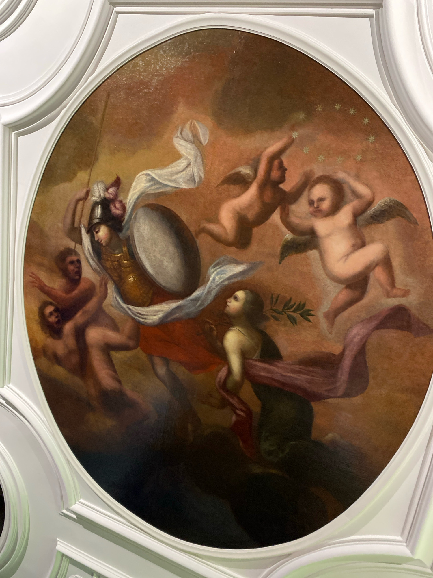 The oval painting depicts a mythological or allegorical scene featuring several winged cherubs and a warrior-like figure. The central figure, possibly Minerva or another goddess of war and wisdom, wears armor, a helmet adorned with feathers, and carries a shield and a spear. She is depicted in motion, surrounded by flowing drapery. Two cherubs are shown in flight, one reaching towards a scattering of stars on the upper right, and another in a dynamic pose. Below and to the left of the central figure, two nude male figures appear to be reaching out or struggling. In the lower right quadrant, a female figure with a green garment and a purple sash is depicted with a laurel branch. The overall color palette is warm, with shades of orange, brown, and pink dominating the background and figures, contrasted with the white of the cherubs' skin and drapery. The painting is set within a white, ornate architectural frame. This artwork is located in the State Hermitage Museum, Saint Petersburg, Russia.