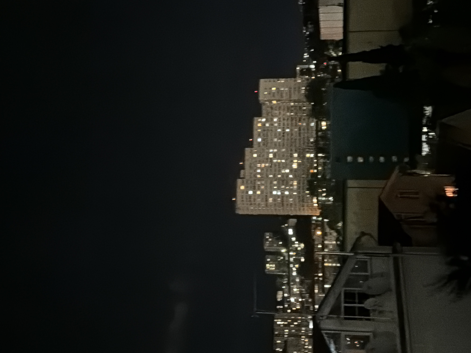 A nighttime aerial view of a city skyline in Sochi, Russia, shows a dense urban landscape dominated by apartment buildings. The buildings are illuminated with countless windows, creating a warm, speckled pattern against the dark sky. In the foreground, smaller buildings and structures are visible, with some also showing interior lights. The sky is mostly dark, with a faint cloud formation on the left side. The scene appears to be a typical residential area at night, characterized by its lit-up buildings and dark sky.