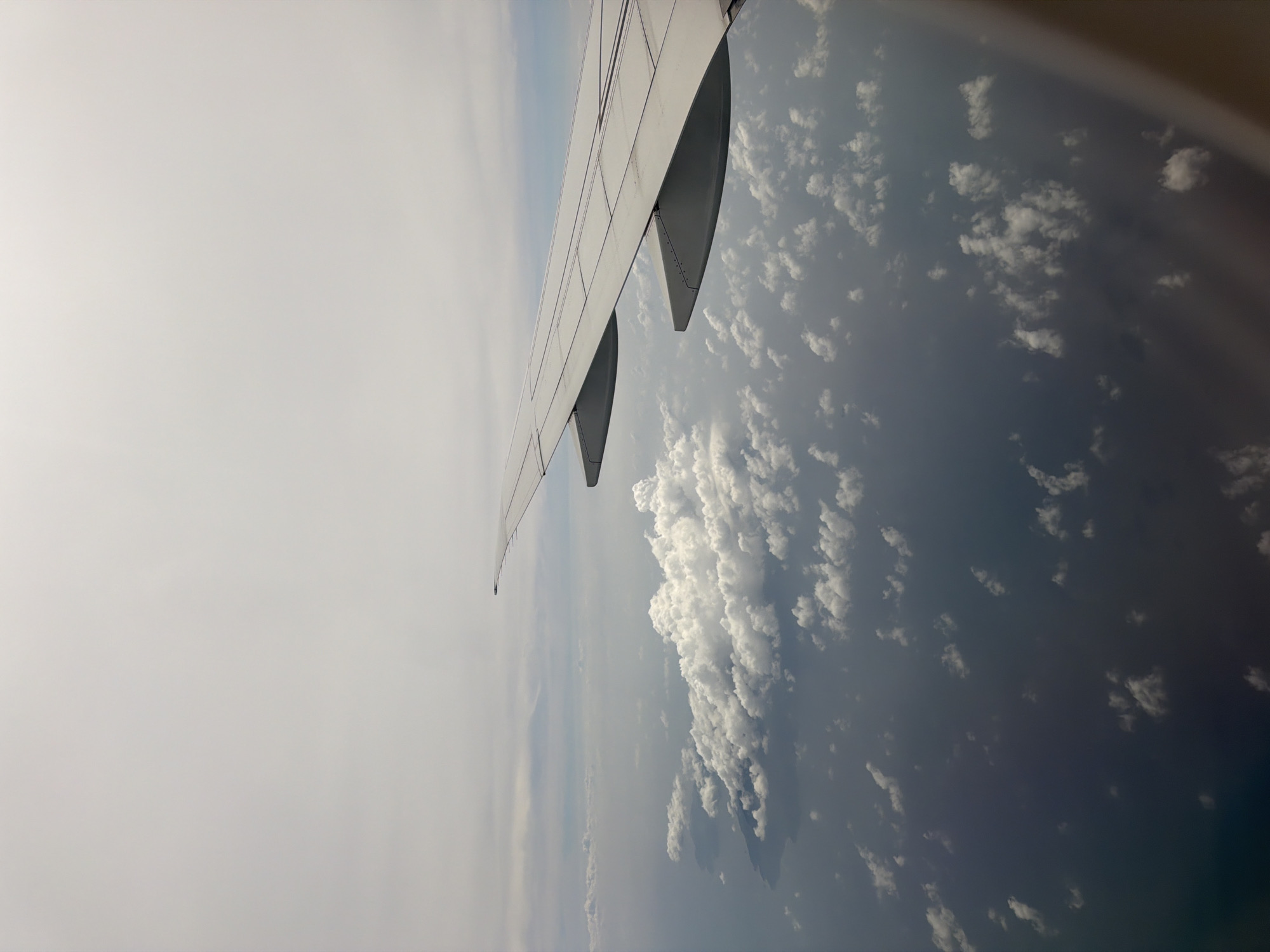 An aerial view from an airplane window shows the wing of the aircraft extending into the frame from the right, with a portion of the ocean and clouds below. The ocean appears dark and vast, with scattered white clouds reflecting light on its surface. The clouds are varied in size and density, with some appearing fluffy and well-defined while others are more diffuse. A faint landmass can be seen in the distance on the left side of the image, partially obscured by clouds. The sky above is overcast with a light, diffused light, suggesting a cloudy or hazy day. The image captures a serene and expansive scene from high above. There is no indication of the city of Singapore in this image.