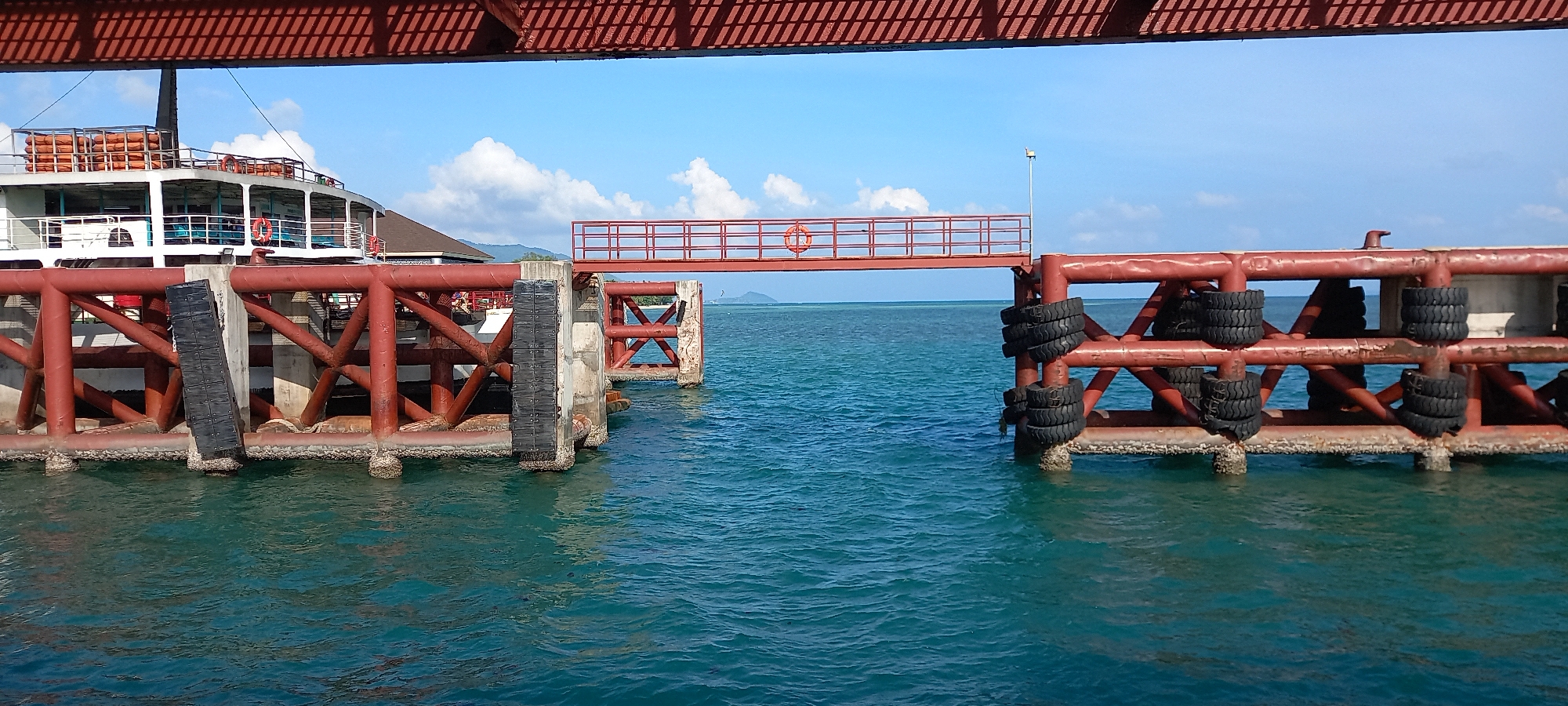 This image depicts a sunny daytime scene at a pier or ferry terminal, identified by the location context as Thong Sala, Thailand.
The primary subjects are a robust red metal pier structure and a large white ferry. The pier is constructed with thick red beams and supported by concrete pilings. Numerous black car tires are attached to the pier's side, serving as fenders to protect docking vessels. On the left side of the image, the bow and midsection of a multi-deck white ferry or boat are visible, docked alongside the pier. Orange life vests are stacked neatly on its upper deck, and some seating can be seen through its windows.
A separate red metal gangway with railings extends horizontally across a gap in the pier structure, seemingly connecting two sections or providing access to another area. A red lifebuoy is attached to this gangway's railing, and a lamppost stands near its far end.
The water is a clear, vibrant blue-green, showing ripples from gentle movement. In the background, a bright blue sky with scattered white clouds dominates, and distant green land, likely an island or coastline, is visible on the horizon. The strong sunlight casts prominent shadows from the overhead structure onto the water, indicating clear and bright weather.
No discernible text is clearly visible on any objects in the image. The scene suggests typical maritime activity at a coastal port, with a ferry either docked or preparing to depart/arrive.