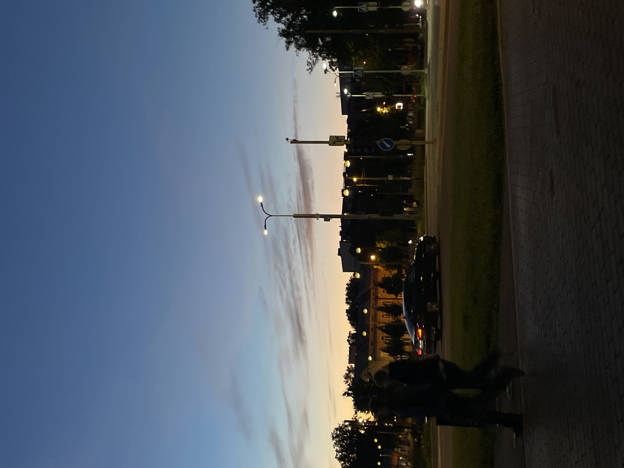 The image depicts a street scene in Tallinn, Estonia at dusk. The sky is a gradient of deep blue to pale orange, with wispy clouds. Two individuals are walking on a brick sidewalk in the foreground, silhouetted against the fading light. Behind them, a dark-colored car is driving on the road, its taillights illuminated. Several streetlights with bright yellow bulbs are visible, illuminating parts of the street and sidewalk. In the background, buildings and trees line the street, with more streetlights adding to the ambiance. The overall impression is a calm evening in a city environment.