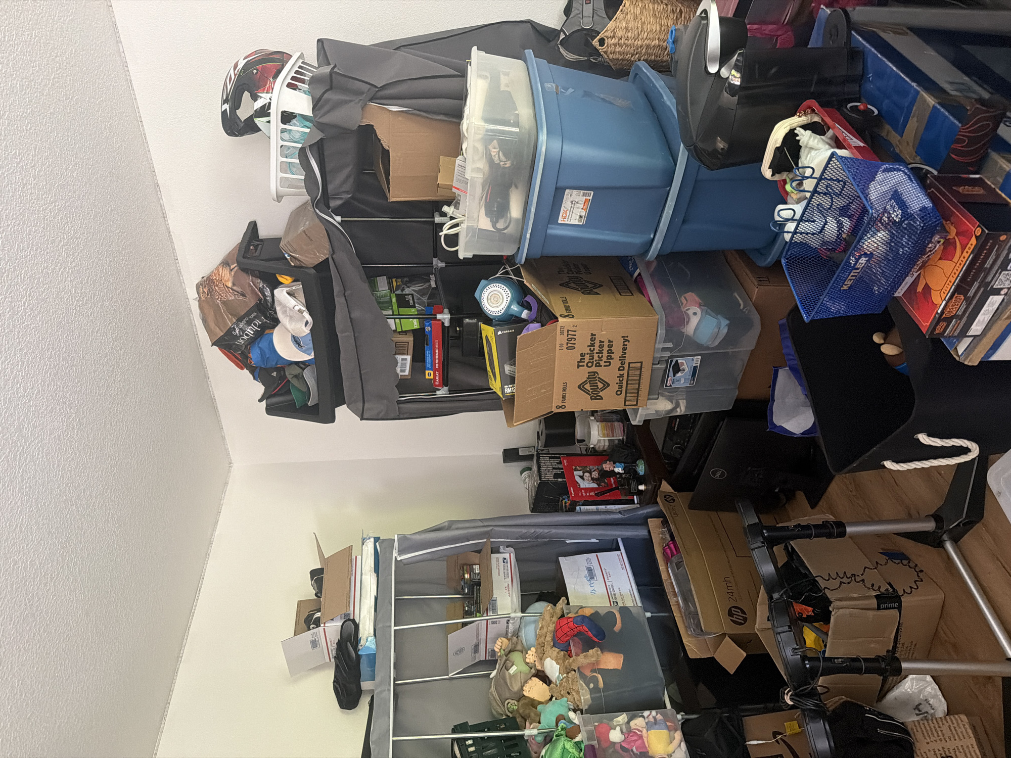 The image shows a cluttered room, likely a storage area or a room undergoing organization. The scene is filled with various boxes, plastic bins, stuffed animals, and other household items. There is a tall, gray fabric-covered shelving unit on the left, filled with boxes and stuffed animals. To the right, a black fabric-covered storage unit is packed with items, including a red and black helmet on top. Various boxes are stacked on the floor and on shelves, including a large brown box from Bounty. A black Keurig coffee maker is visible on top of blue plastic bins. The room appears to be indoors, with white walls and a textured ceiling. The floor is a light-colored wood. There are no people visible in the image. The overall impression is one of disarray and storage.