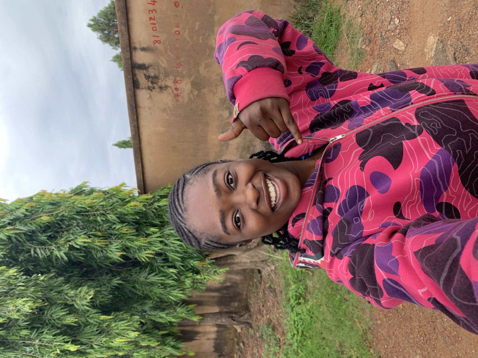 An image taken in Rafin Par, Nigeria, features a young woman with dark skin and braided hair, smiling broadly directly at the camera. She wears a pink, purple, and dark grey abstract camouflage-patterned hooded sweatshirt with a silver-colored zipper. Her right hand is raised, forming a "shaka" gesture with the thumb and pinky extended. The background on the left is dominated by a large tree with dense, drooping green foliage and visible trunks, with green grass and earthy patches at its base. A weathered, light brown wall spans the mid-ground, exhibiting dark stains and red numbers written on it, specifically "8101234" on the upper right and "07084400" on the lower middle. Above the wall, a cloudy, light grey sky is visible, with sparse green trees in the distant background. The foreground ground consists of reddish-brown dirt or gravel with minimal scattered debris.
