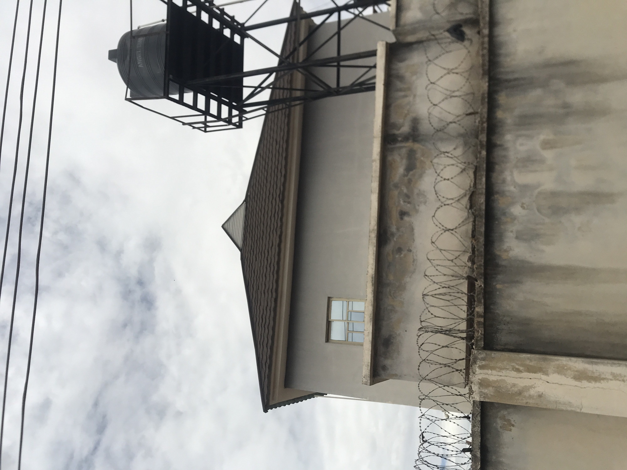 This image captures an exterior view of a property in Rafin Par, Nigeria, under an overcast sky marked by gray and white clouds and three dark power lines spanning the upper frame. In the foreground, a weathered concrete perimeter wall, exhibiting stains and cracks, is topped with multiple coils of razor wire acting as a security barrier. Behind this wall, a light gray building with a sloped brown corrugated metal roof is partially visible. A rectangular window with multiple panes is located on the building's side, above the wall. To the right, a tall, black metal lattice structure supports a large, dark gray plastic water tank, upon which the text "ANGEL TANK" is discernible. No individuals are present in the scene.