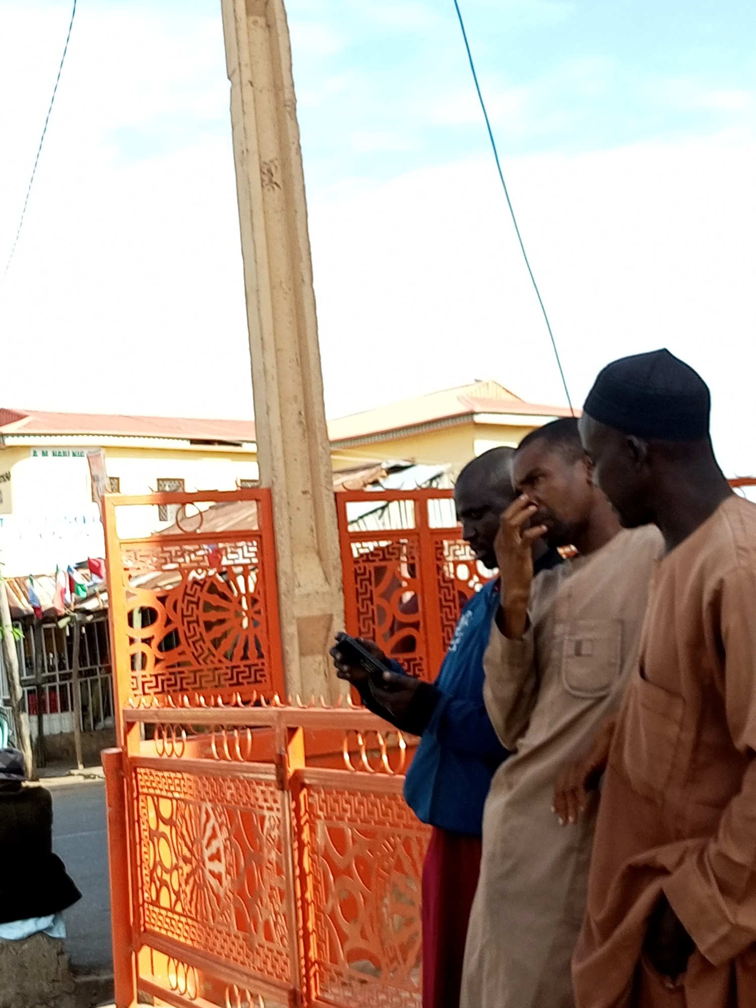 The image depicts three men standing outdoors in Arawa, Nigeria. The men are dressed in traditional attire, with two wearing kaftans and one wearing a blue tunic. The man in the middle is touching his face, and the man on the right is looking down at a mobile phone. They are standing in front of an orange metal gate with intricate patterns. In the background, a building with a red roof and yellow walls is visible. A utility pole with a blue wire extending from it is also present in the scene. The sky is partly cloudy.