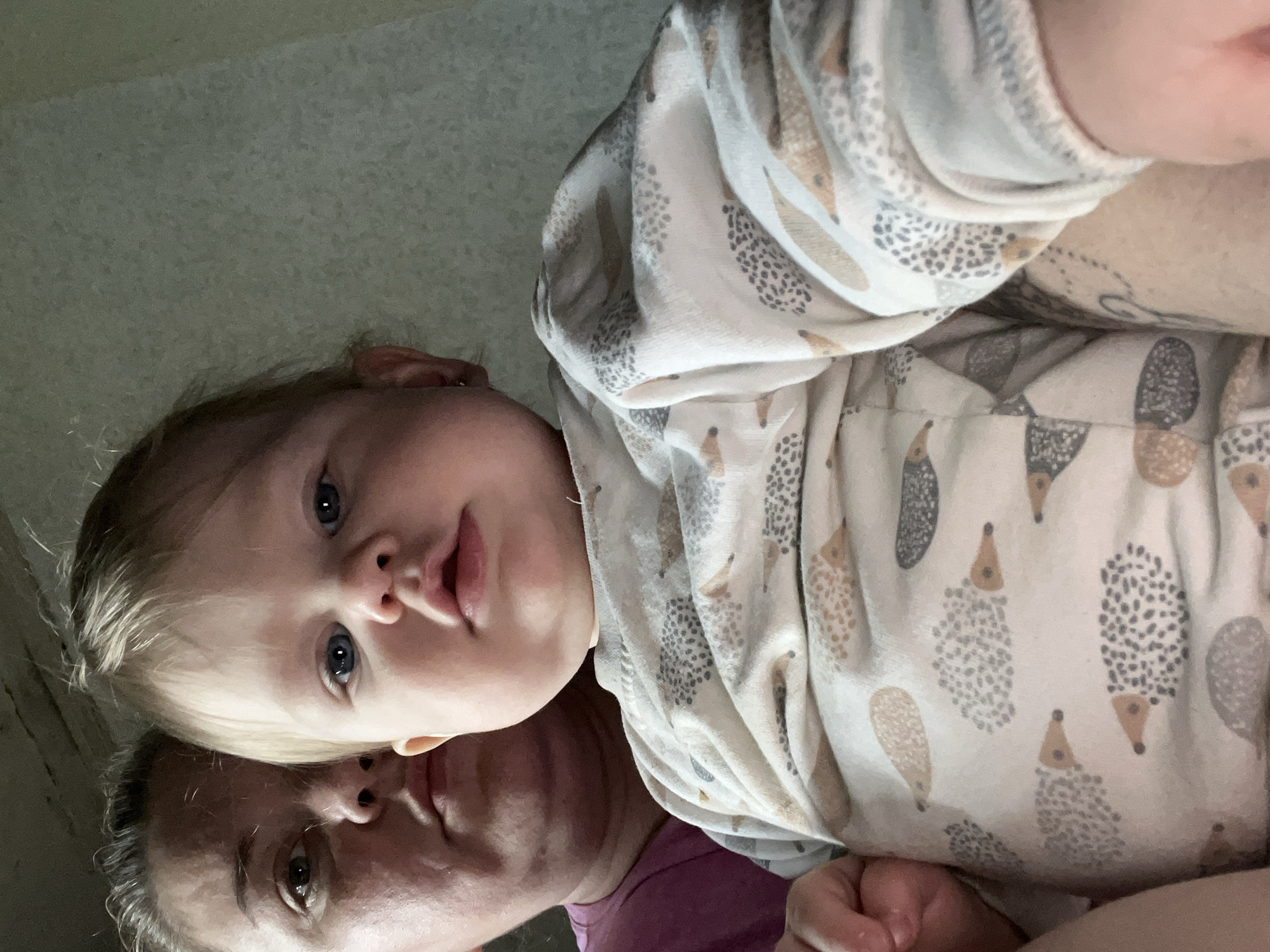 The image, oriented vertically, captures a close-up, intimate portrait of two individuals indoors, likely in Фоки, Russia. A young child, possibly a toddler, with light blonde hair and bright blue eyes, is positioned in the upper portion of the frame. The child looks directly towards the camera with a neutral expression and slightly parted lips. They are wearing a light-colored top adorned with a repeating pattern of cartoon hedgehogs in shades of grey and light brown.
Below the child, an adult's face is partially visible, oriented horizontally across the lower part of the frame. The adult, appearing to be female with light hair, is seen from the nose down, looking slightly towards the left side of the child. She is wearing a purple or magenta top, partially visible on the right side of the frame.
The lighting is soft and somewhat dim, suggesting an indoor environment without direct harsh light. The background is a plain, light grey wall, which is out of focus, indicating a shallow depth of field. The composition suggests a close-up photograph, emphasizing the connection between the child and the adult, with the unusual vertical orientation possibly indicating an accidental rotation or a specific photographic style. No visible text is present in the image.