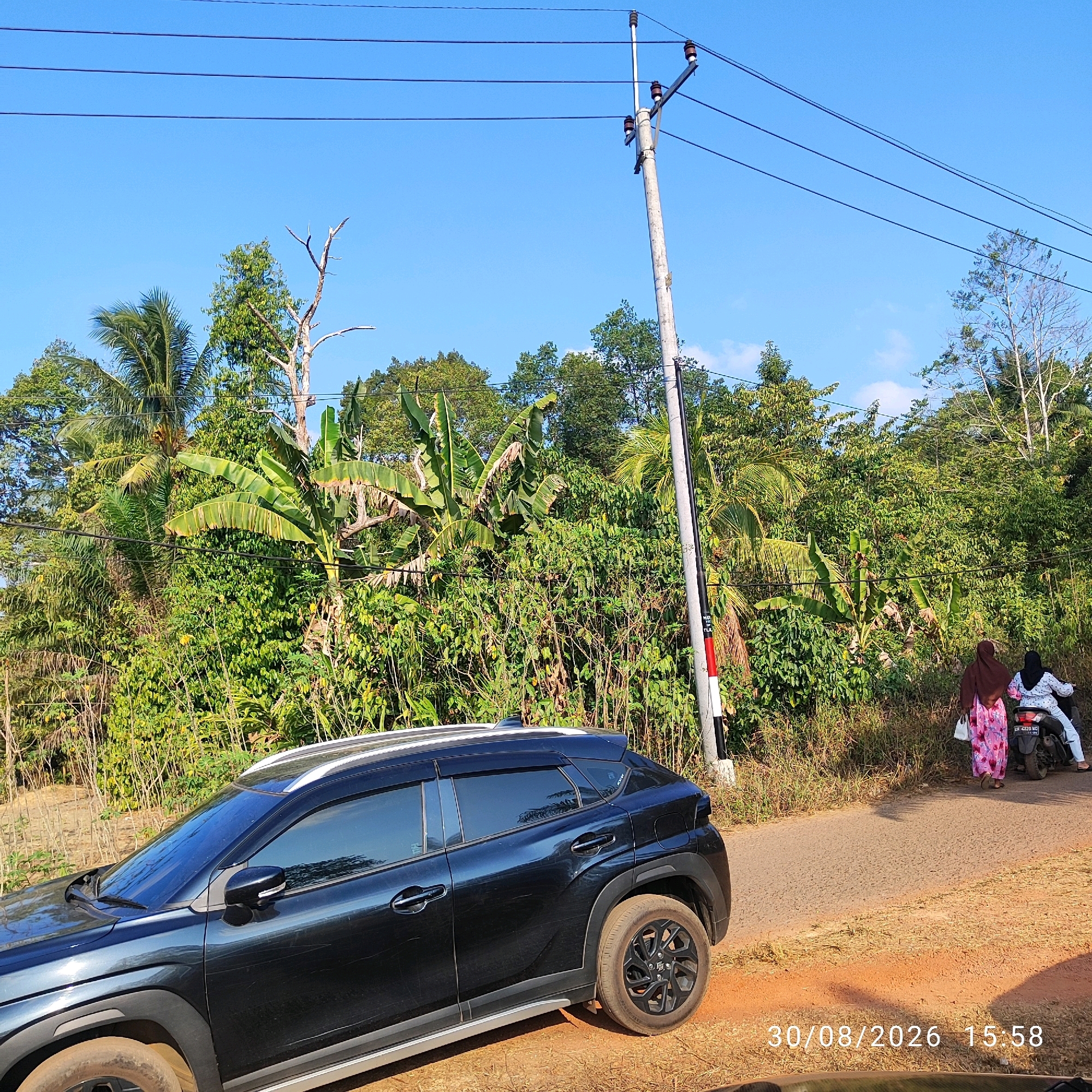 The image depicts a rural scene in Pembaharuan, Indonesia, on August 30, 2026, at 15:58. A black SUV is parked on the left side of a paved road, with its front half visible. The road extends into the background, where two individuals are seen on a motorcycle, one driving and the other as a passenger, both wearing headscarves. The landscape is dominated by lush, dense green vegetation, including banana trees and palm trees, indicative of a tropical environment. A utility pole with power lines runs across the upper portion of the frame, contributing to the rural setting. The sky is clear and blue. The ground is a mix of dry grass and reddish-brown soil, particularly noticeable around the parked vehicle.