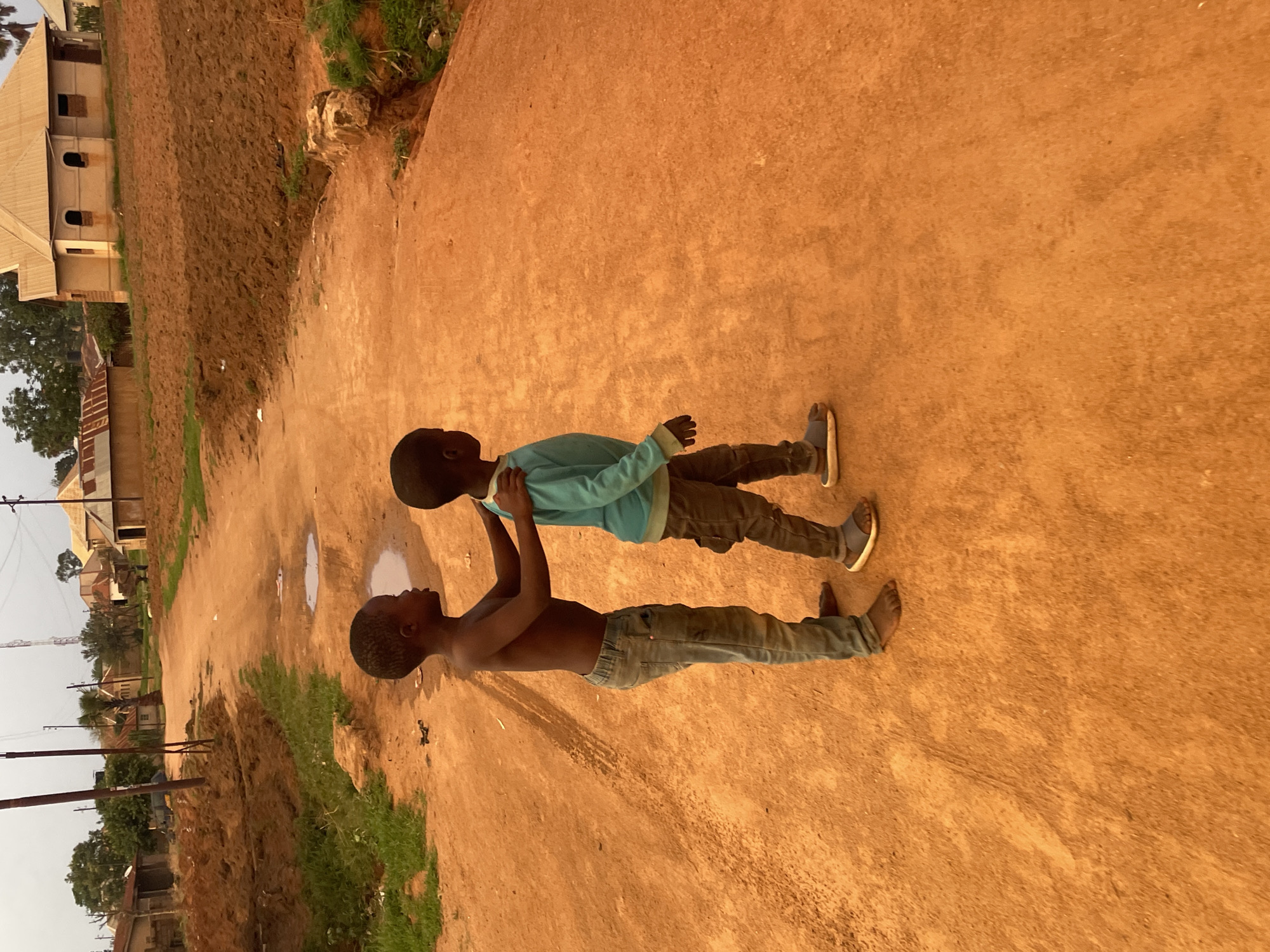 Two dark-skinned boys are standing on a dirt road in Bukuru, Nigeria. The boy closer to the camera is shirtless with denim jeans. The other boy is wearing a long-sleeved blue shirt and gray pants with sandals. The background shows modest buildings, trees, and dirt fields. Power lines are visible. There is a puddle on the road behind the boys.