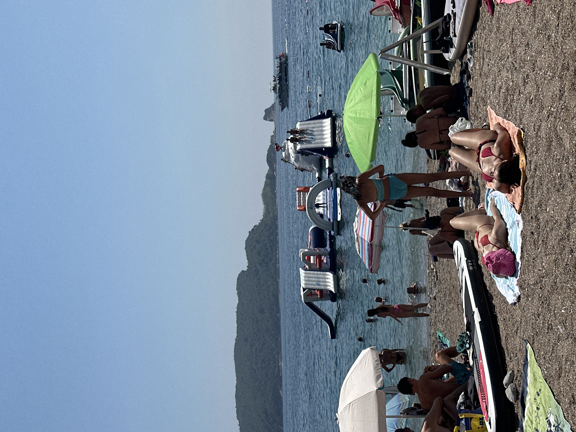 The image depicts a crowded beach scene in Budva, Montenegro, on a clear, sunny day. The sea is calm and blue, with a chain of inflatable water slides and obstacles positioned offshore. Several people are enjoying the water, swimming, and interacting with the inflatable attractions. A boat with passengers is visible in the background, sailing on the sea. On the pebbled beach in the foreground, various individuals are sunbathing, sitting under umbrellas, and relaxing. A paddleboard lies horizontally on the beach, with a yellow bag nearby. The overall atmosphere suggests a lively and recreational summer day at the seaside.