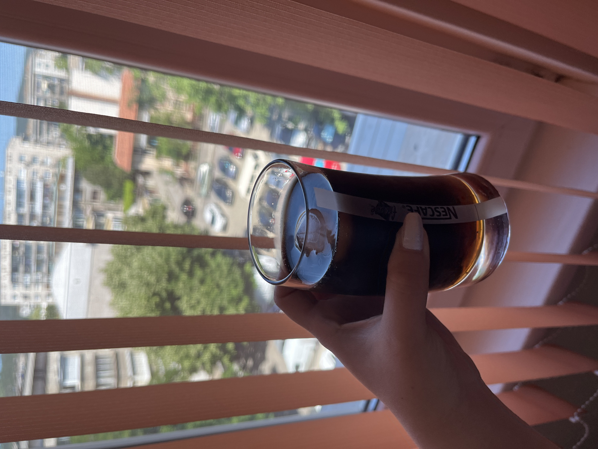 A person's hand, with neatly manicured nails, holds a glass of iced coffee in front of a window with beige vertical blinds. The glass has a white label that reads "NESCAFE". Beyond the blinds, a city scene is visible, featuring apartment buildings, trees, and parked cars. The buildings appear to be from the socialist realism architectural style, common in Bulgarian cities like Stara Zagora. The lighting suggests it is daytime, possibly late morning or afternoon.