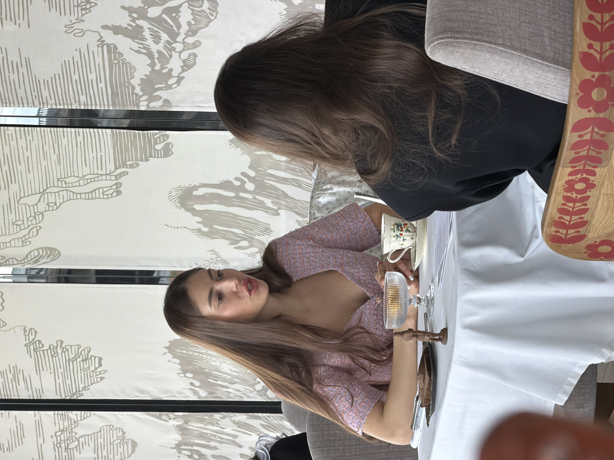 Two women are seated at a table in a restaurant. The woman on the left is wearing a light purple patterned dress. She is talking and looking towards the other woman. There is a glass of water in front of her, and a teacup and saucer on the table. The other woman is seen from the back, with long brown hair and wearing a black shirt. The table is covered with a white tablecloth, and there is a decorative wooden item with floral motifs on the table. The background is a patterned wallpaper. While location cannot be confirmed with certainty based solely on the image, the description provided does not fit.