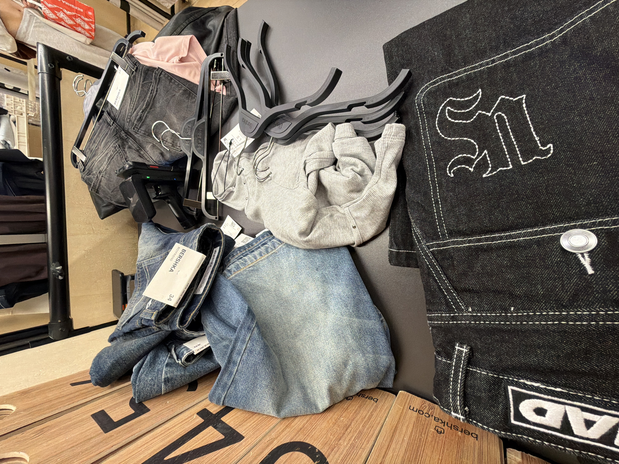 The image shows a display of clothing items in what appears to be a retail store, likely a Bershka outlet, given the brand's labeling on the items and tags. The clothing includes jeans in various shades of denim, ranging from light blue to black, along with a grey sweater and a pink shirt. All items are hanging on hangers. The items are situated on a wooden surface, possibly a display table, which bears the Bershka logo and website address "bershka.com". The size indicators of the clothes are visible, implying the clothes are for sale, indicating a store or storage. The exact location of the image in the city of Sofia, Bulgaria, is not specified.