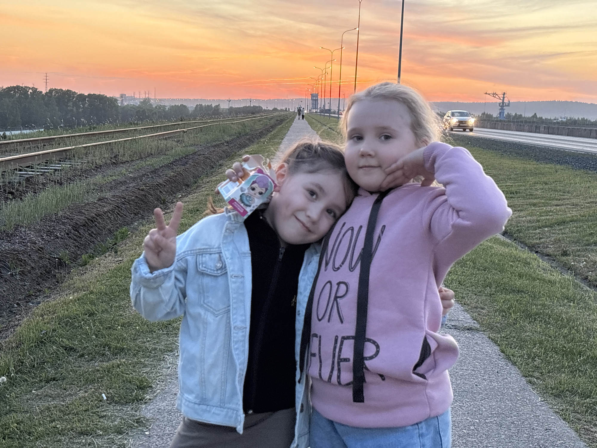 Two young girls are posing for a photo. The girl on the left is holding up a peace sign and holding a small toy box. The girl on the right is holding the other girl’s face. They are standing on a paved pathway. To the left of the pathway are railroad tracks with grass growing between them. To the right of the pathway is a road with a car driving on it. In the background is a sunset sky. The image was taken in Chaikovsky, Russia.