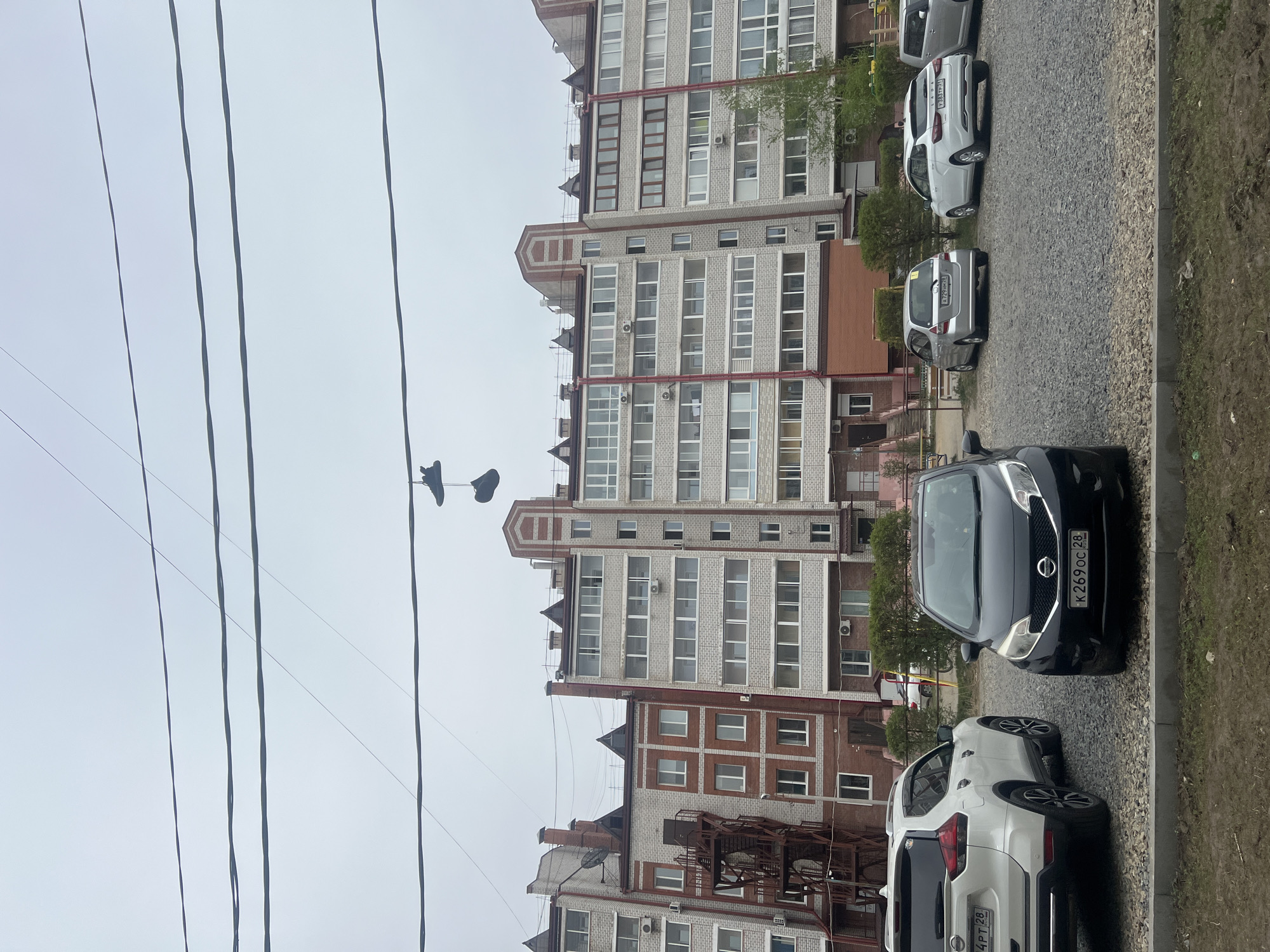 The image depicts an urban scene in Unknown, Unknown, showing a residential building with multiple stories, constructed primarily of light gray bricks with red-brown brick accents around the windows and edges. A series of power lines run across the upper left portion of the frame, and a pair of shoes hangs suspended from one of the wires. Along the bottom right edge of the image, several parked cars are visible, including a black Nissan with the license plate "K269OC 28" and a white SUV with the license plate "B444PT 28." The ground is covered with small rocks and some patches of green vegetation along the edge. The sky is overcast.
