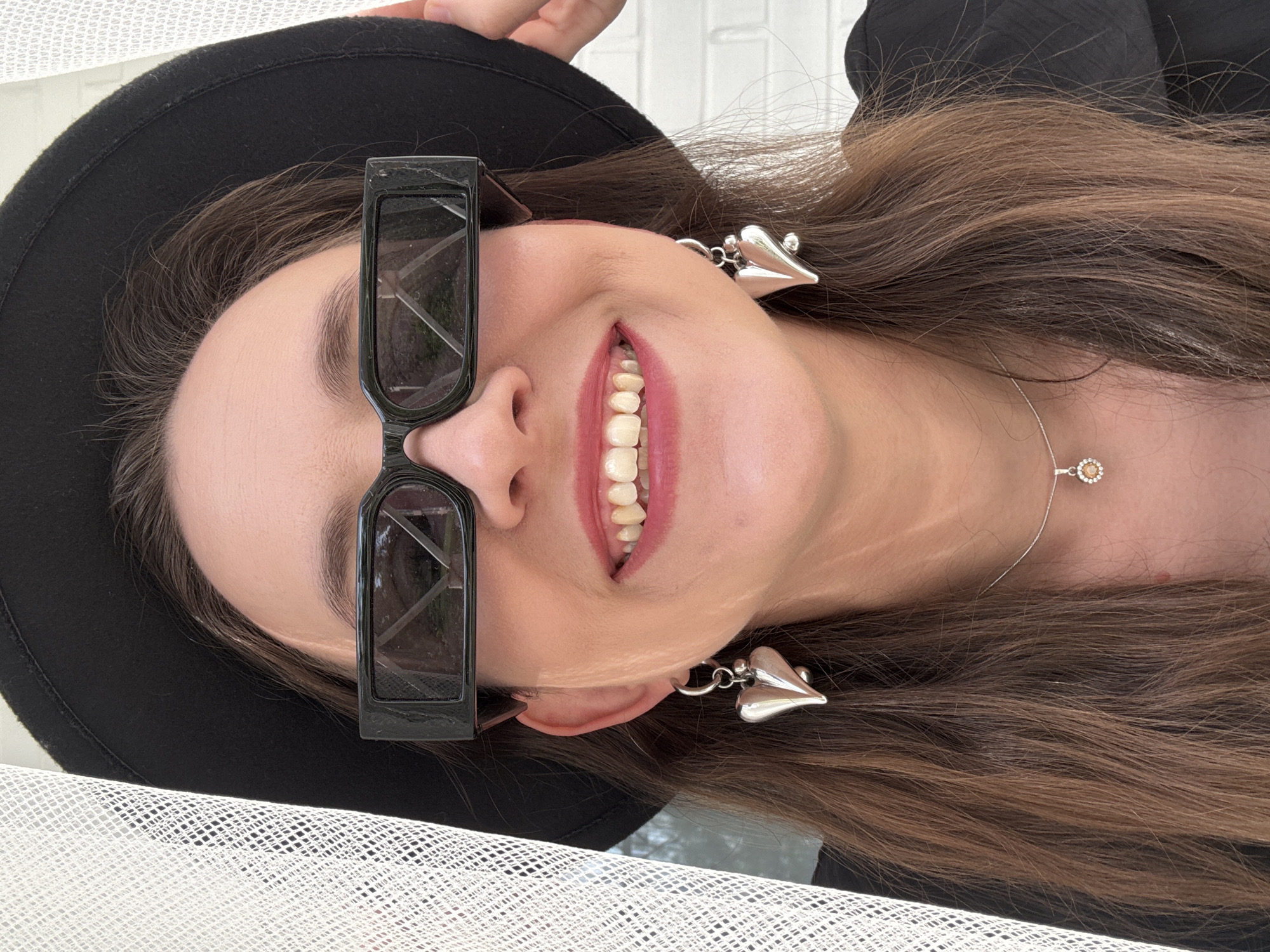 A woman with long brown hair smiles while wearing sunglasses, a black hat, and heart-shaped earrings. She is wearing a silver necklace. The background includes a white brick wall and white netting. It is not possible to confirm the location as Chaikovsky, Russia from the information provided.