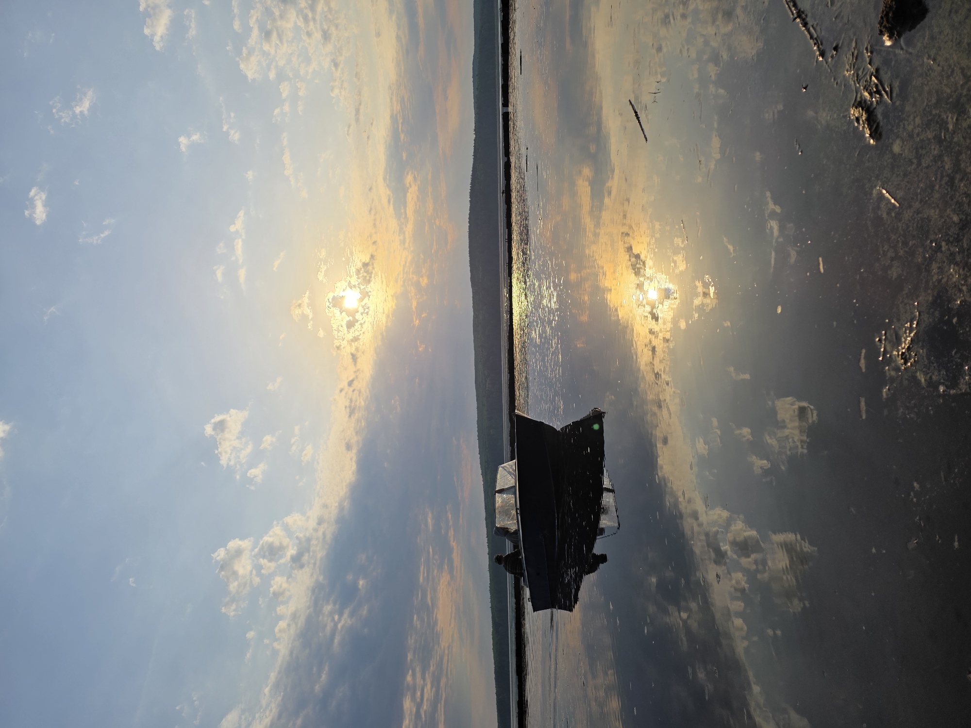 The image shows a boat on a body of water, likely a lake or river. The sky is visible above, with clouds and the sun shining through. The reflection of the sky and sun can be seen in the water. There is a silhouette of land in the distance. The boat appears to be a small, metal fishing boat with a person inside. There's some debris in the water near the shore. It's a calm, possibly sunset or sunrise scene. The image is rotated 90 degrees counter-clockwise. The location is specified as the city of Chaikovsky, Russia, indicating this is where the photograph was taken.