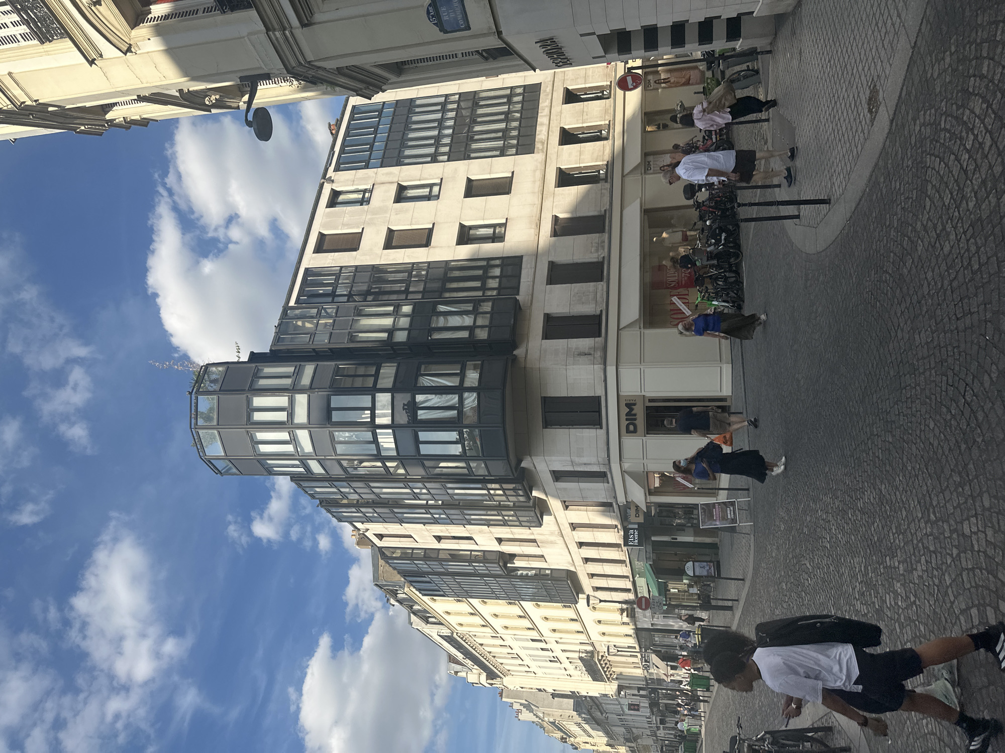 The image depicts a street scene in Paris, France, likely on a sunny day given the bright lighting and clear blue sky with scattered clouds. The street is paved with cobblestones and lined with buildings. The primary building in the center is a modern, multi-story structure with a distinctive curved, glass-enclosed upper section, topped with greenery. This building features a facade with multiple windows, some of which are dark and reflective, and others that are lighter and reveal interior curtains. The ground floor of this building appears to house retail establishments, with signage visible for "DIM" and "ELSA'S HOME".
To the left, a row of older Parisian buildings stretches down the street, characterized by their light-colored facades, numerous windows, and balconies. These buildings also appear to contain shops on their ground floors. To the right, the edge of another building is visible, with a sign for "SEPHORA" and a street sign indicating "15 ARR. RUE DE COMMERCE".
Several people are present on the street. In the foreground, a young Black person wearing a white t-shirt, black shorts, and black and white athletic shoes walks towards the left, carrying a dark jacket over their shoulder. Further down the street, a group of people are walking, some looking at shops. A woman in a blue top and long skirt walks towards the left, another person in a blue top and dark skirt walks towards the right, and a man in a white shirt and dark shorts walks towards the right. There are also bicycles parked in front of some of the shops. The overall impression is a busy urban environment with a mix of modern and older architecture.