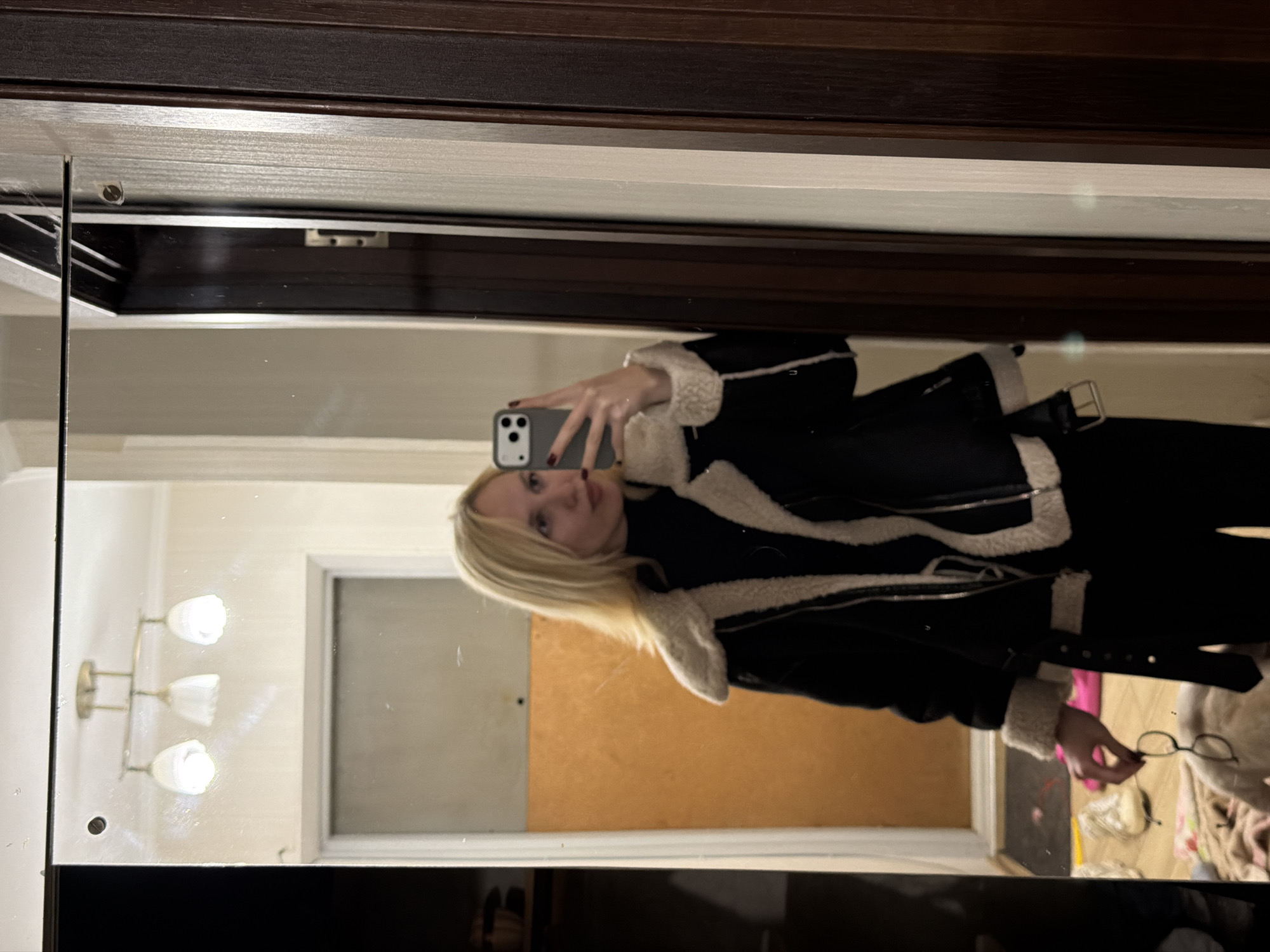 A young woman with blonde hair stands in front of a mirror, taking a selfie with her phone. She is wearing a black jacket with a cream-colored furry lining, a black turtleneck, and black pants. She is holding a pair of glasses in her right hand. In the reflection of the mirror, a light fixture with three lamps is visible on the ceiling, along with a door with wooden panels to her right. To her left, a closet door is partially open, revealing some items on the floor. The overall scene appears to be an indoor setting, possibly a hallway or a room with a mirror. The location is Cheltuitori, Moldova.