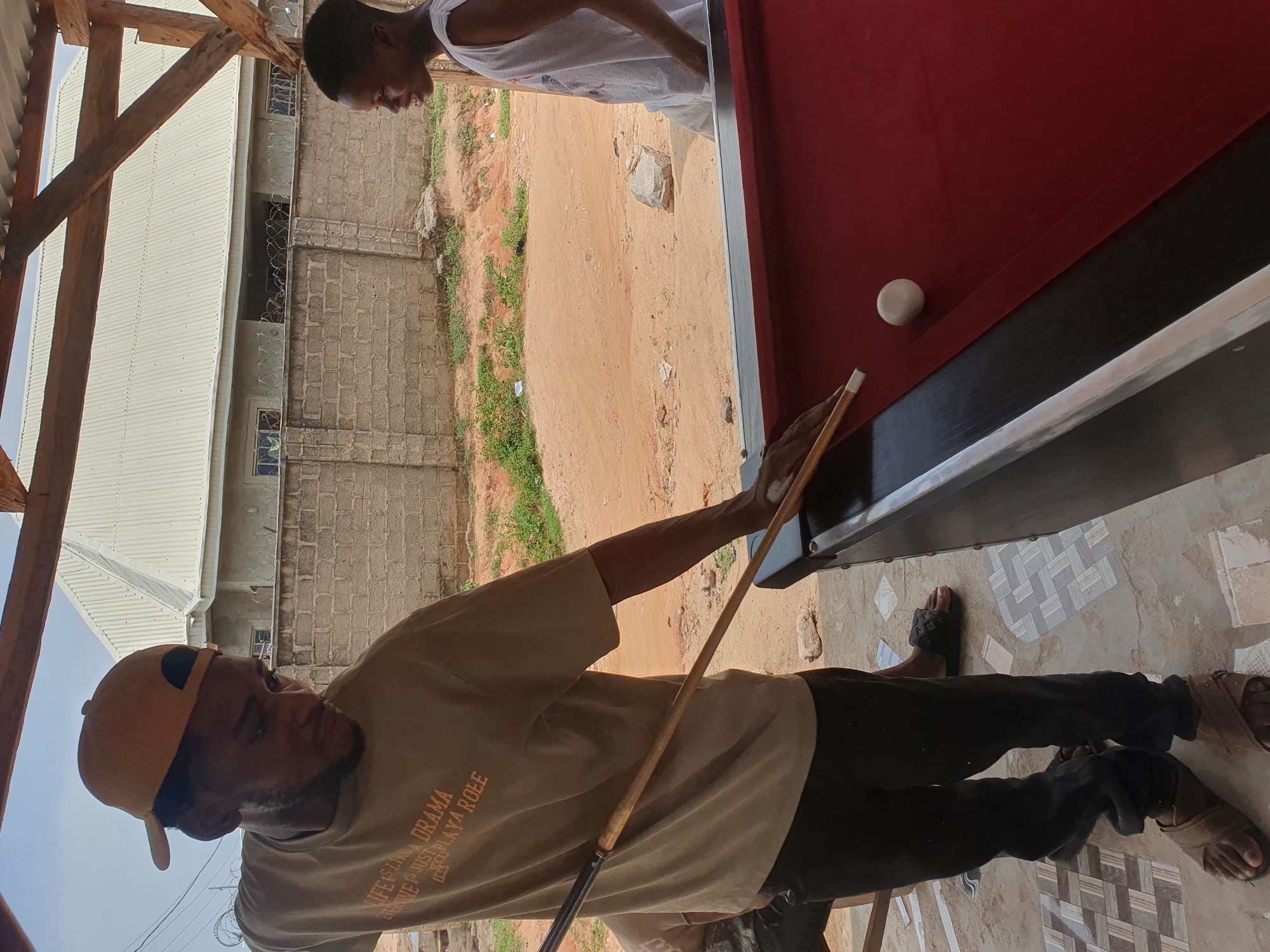 The image depicts two young men at an outdoor pool table in Lokoja, Nigeria. The man in the foreground, wearing a tan t-shirt with orange text and a yellow baseball cap, is actively playing pool, holding a cue stick with his right hand. He is positioned to strike a white ball on the red felt of the pool table. His attire includes black pants and brown sandals.
To his right, a second young man, wearing a white sleeveless shirt, observes the game. He stands facing the table, looking towards the play.
The environment suggests a casual, possibly informal, establishment. A red pool table is the central focus. In the background, a partially constructed building with a corrugated metal roof and a brick wall is visible. The ground is a mix of sand and some patchy vegetation. The overall scene is one of leisure activity in a developing urban setting.