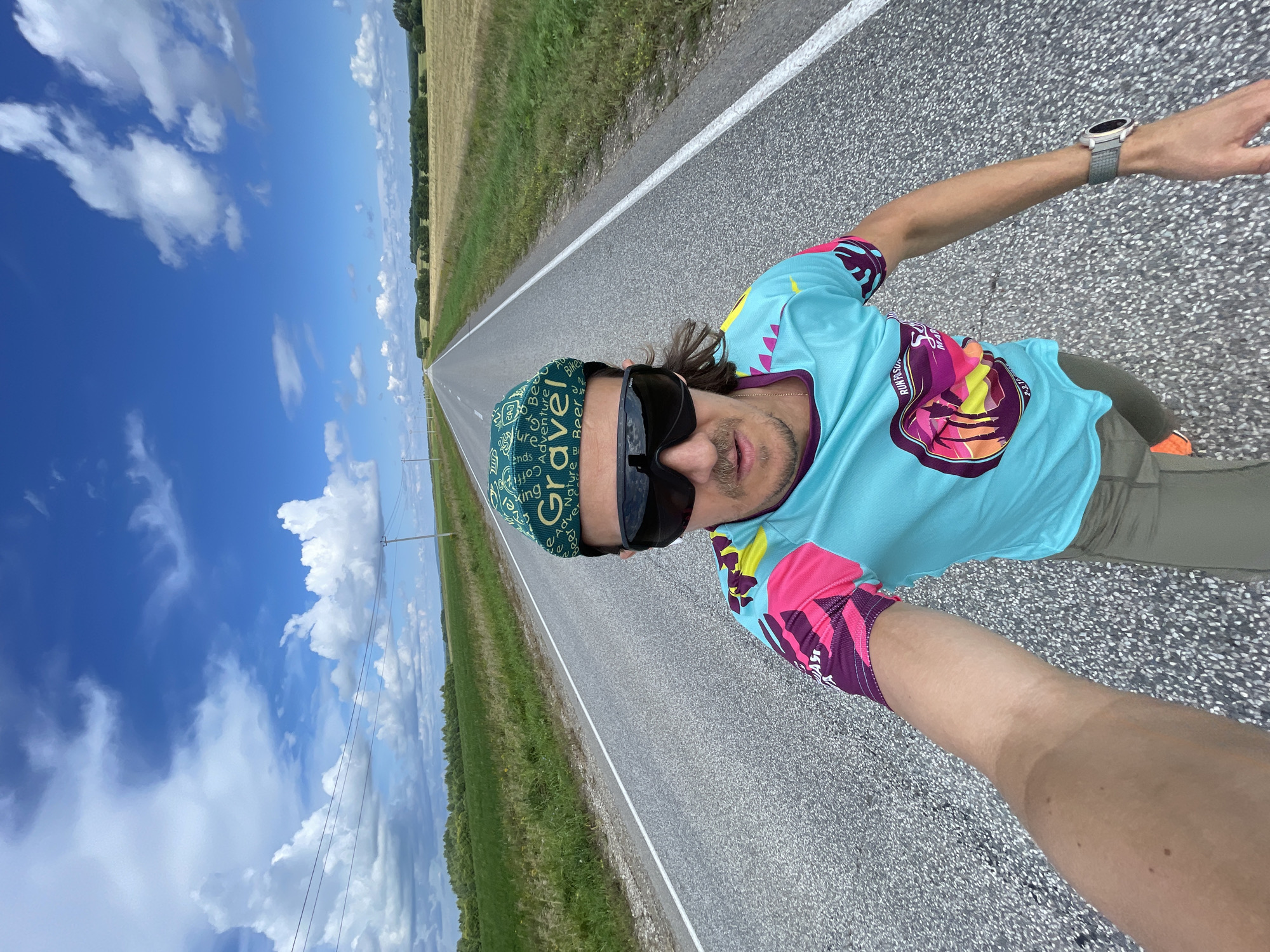 The image is a selfie taken by a man standing on an asphalt road in Perevoznoe, Russia. The man is wearing a teal running shirt with pink and purple accents, a green cap with "Grave Adventure" printed on it, and dark sunglasses. He has a mustache and shoulder-length brown hair. He is wearing khaki shorts and orange running shoes. A smartwatch is visible on his left wrist. The road stretches into the distance, flanked by green fields and a line of trees. The sky is blue with white, puffy clouds. The overall scene depicts an outdoor activity in a rural setting.