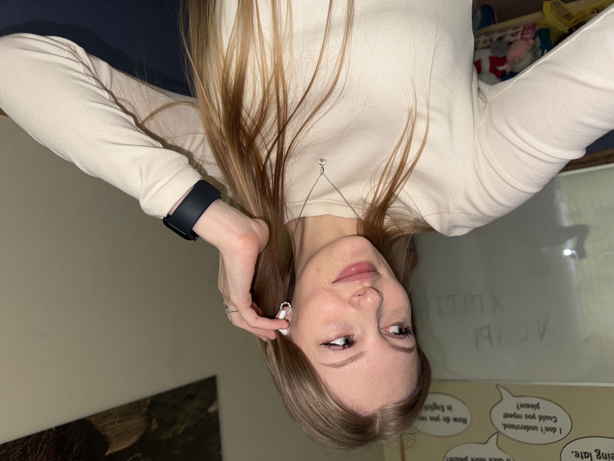 The image is an upside-down selfie of a woman with long, light brown hair wearing a light-colored long-sleeved shirt and a black smartwatch. She is holding a white wireless earbud near her ear with her left hand, which also has a ring on one of the fingers. She is wearing a pendant necklace. In the background, there is a whiteboard with partially erased text written on it in what appears to be Russian ("A ЛОУЫ"). There are also speech bubble-shaped stickers with English phrases such as "I don't understand. Could you repeat please?" and "How do you say..."
It's impossible to determine the location of the city of Perm, Russia, as the image doesn't contain any clues or details that would make it possible to determine the location of the photo.