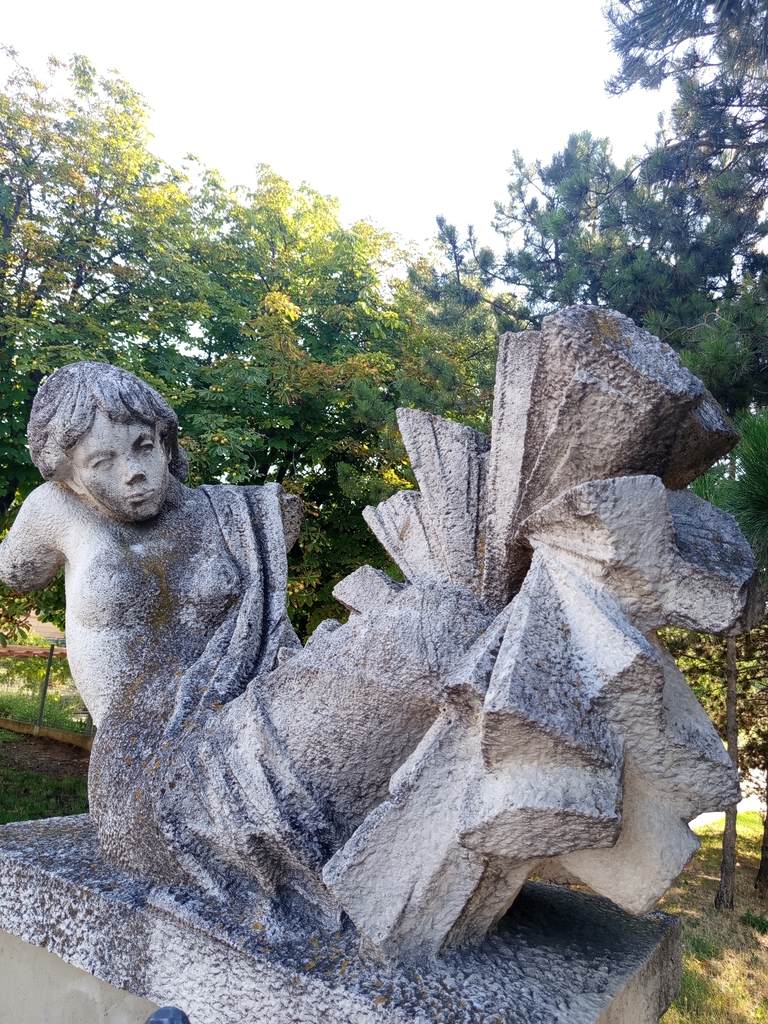 A weathered stone sculpture depicts a reclining figure with a draped garment. The figure's left arm and right shoulder are missing, and its head is tilted to the right, with its gaze directed downwards and slightly to the left. The sculpture is positioned outdoors, with lush green trees in the background. To the right of the figure, a separate abstract stone element features sharp, angular planes. This sculpture is located in Brno, Czechia, specifically in the area of the former Brno Zoo, which is now part of the Lužánky Park.